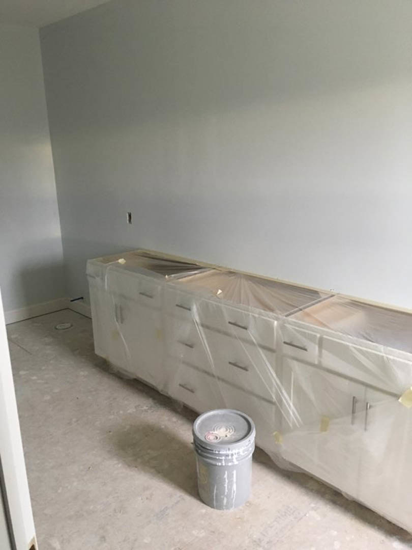 White cabinet partially covered with clear plastic, grey bucket placed in front, tiled bathroom floor and wall visible, window in background.
