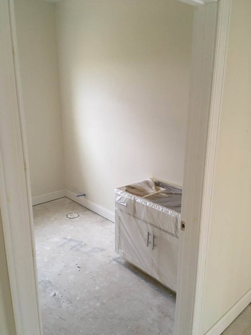 White tile bathroom with built-in sink, white cabinetry wrapped in protective plastic, metal faucet, and smooth plaster walls illuminated by overhead lighting.