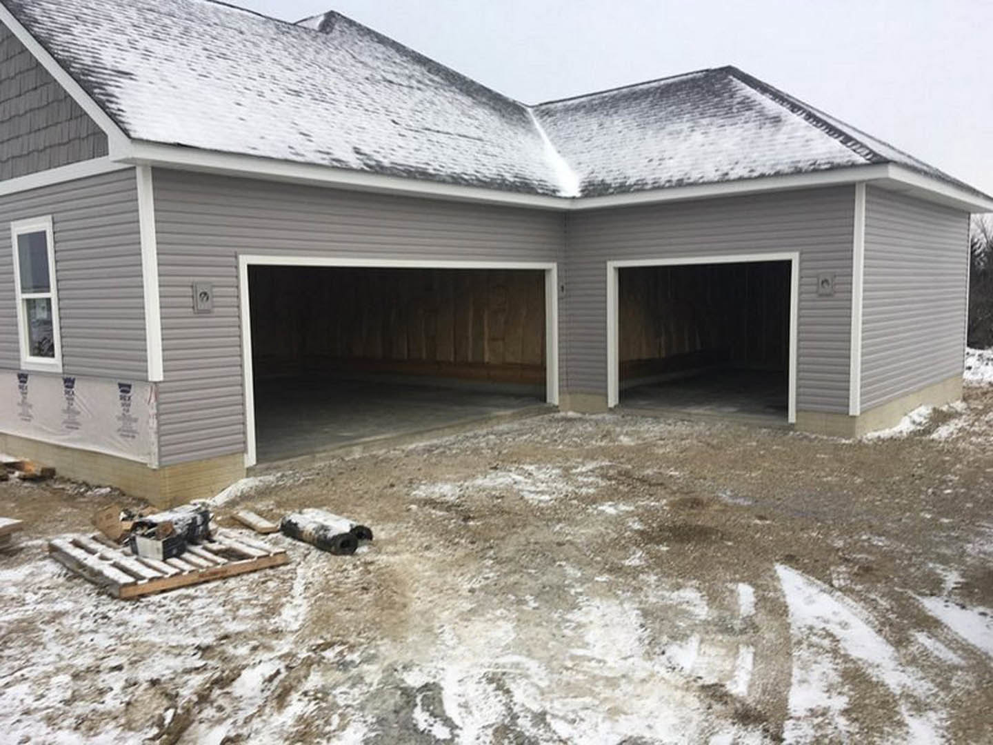 White-framed garage door open to reveal interior, snow covering driveway, roll of carpet near entrance, window on siding, overcast sky above