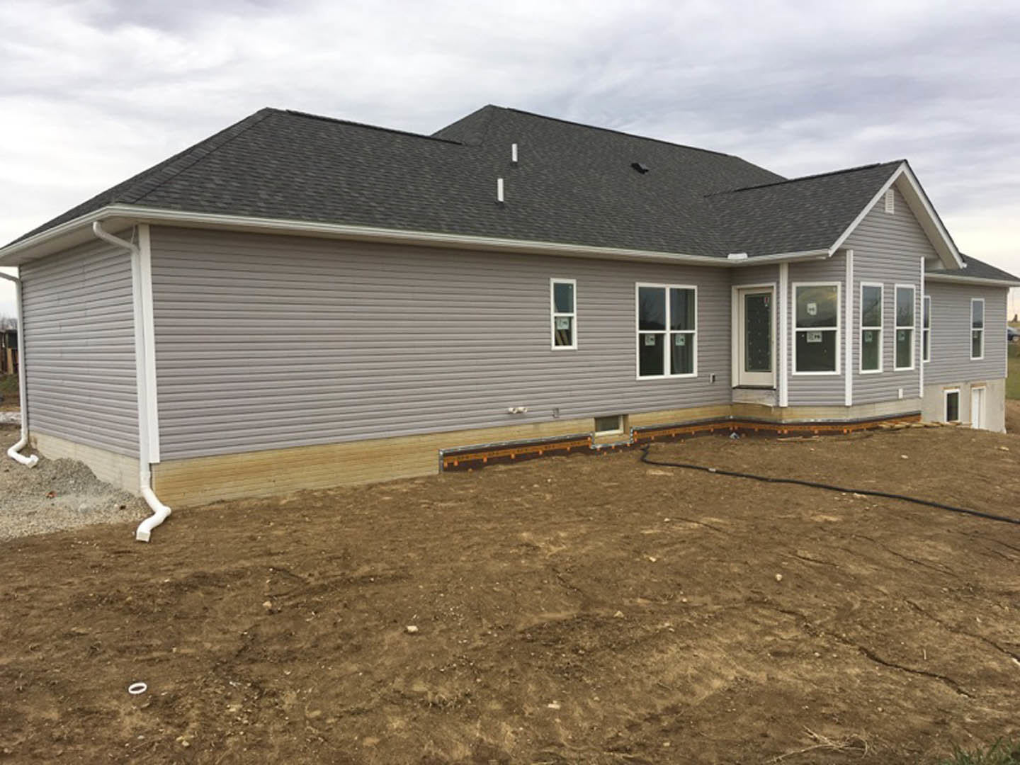 Partially built house with white-framed window displaying a sign, gray siding, shingled roof, exposed dirt yard, and garden hose on the ground under cloudy sky