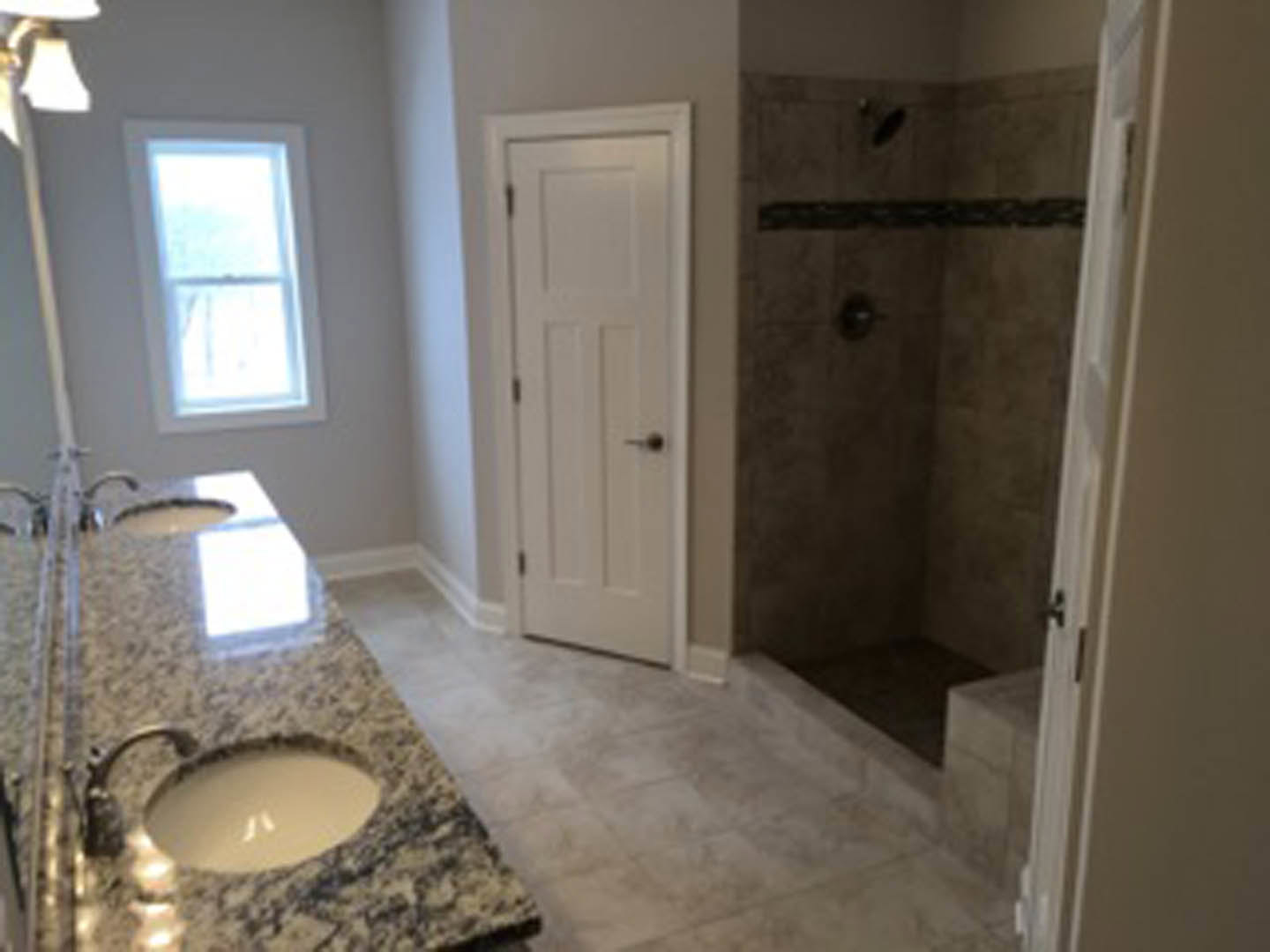 Bathroom featuring marble countertops, undermount sink, glass-enclosed shower with tile walls, white door with black handle, large mirror, and window providing natural light