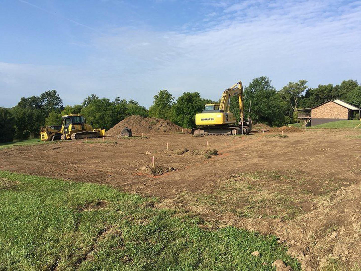 Construction site with yellow excavators and bulldozer on dirt field, partially built house with shingled roof and grassy yard, blue sky with scattered clouds, surrounding trees