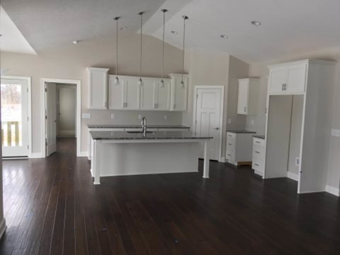 Spacious kitchen featuring a large central island with stone countertop, dark wood flooring, white cabinetry, stainless steel sink, mirrored closet door, and white-framed window.