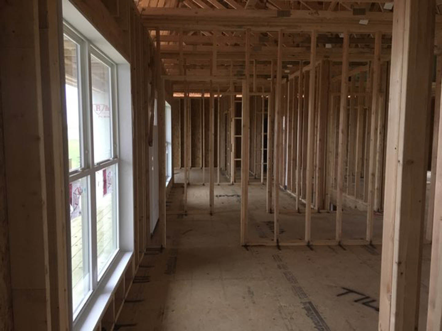 Wood-framed room with exposed beams, large windows with white frames, unfinished plaster walls, and natural daylight illuminating the space.