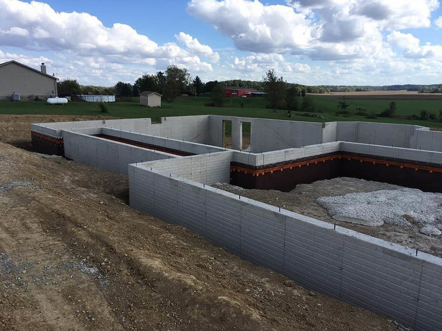 Partially built house with concrete foundation, exposed framing, chimney, and windows, surrounded by dirt field, grass, and trees under a blue sky with clouds.
