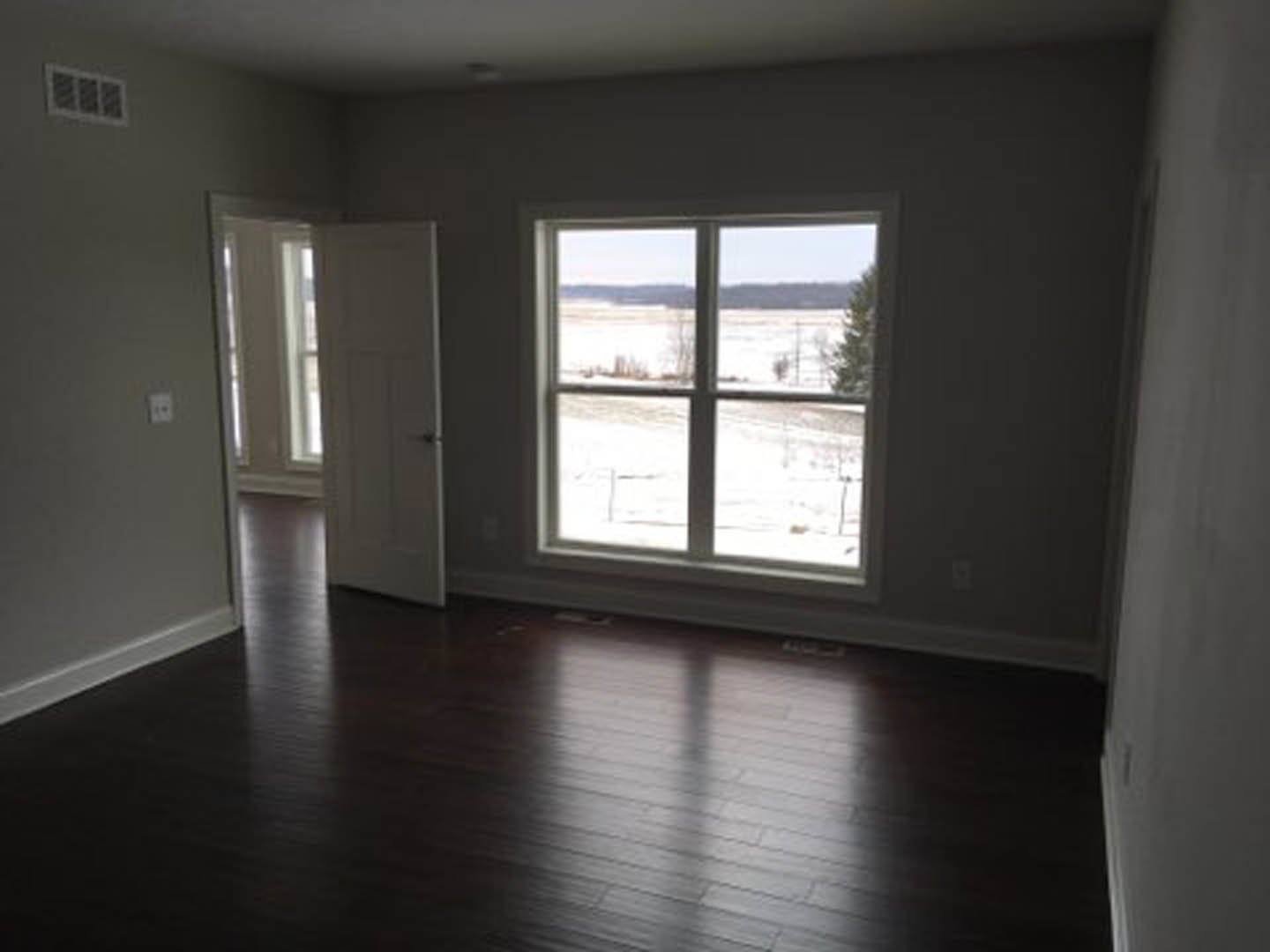 Room with dark hardwood floors, white walls, large window overlooking snowy field, white door with sunlight streaming through, floor vent beneath window
