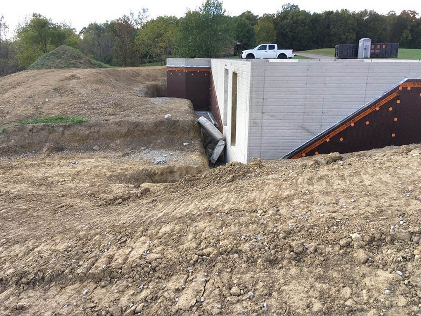 Excavated dirt foundation with concrete ramp, white portable toilet, white truck and car parked near partially built structure, grassy area and trees in background