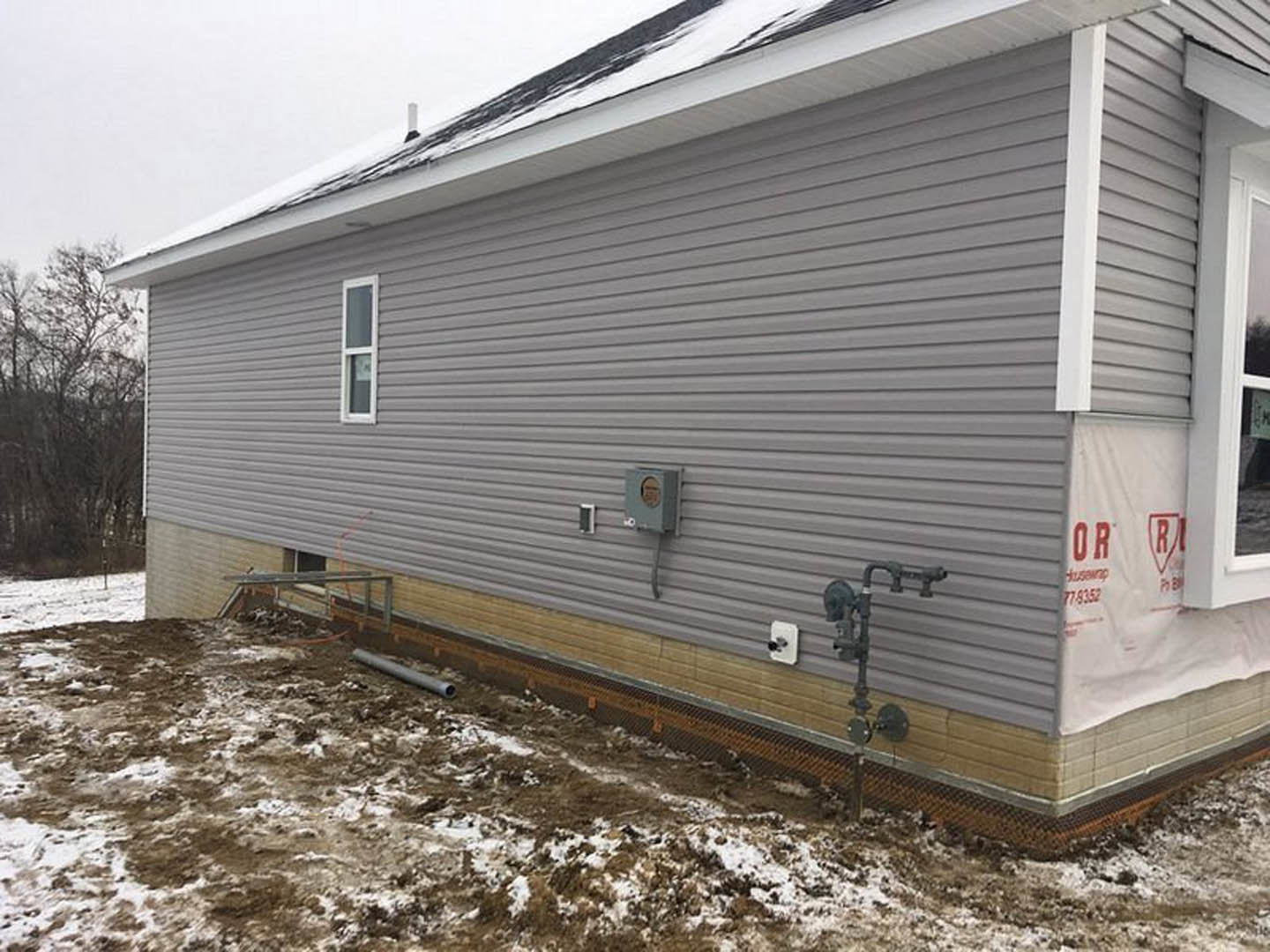 White-walled house with grey siding, white sign featuring red lettering, snow-covered ground, leafless trees in background, overcast winter sky