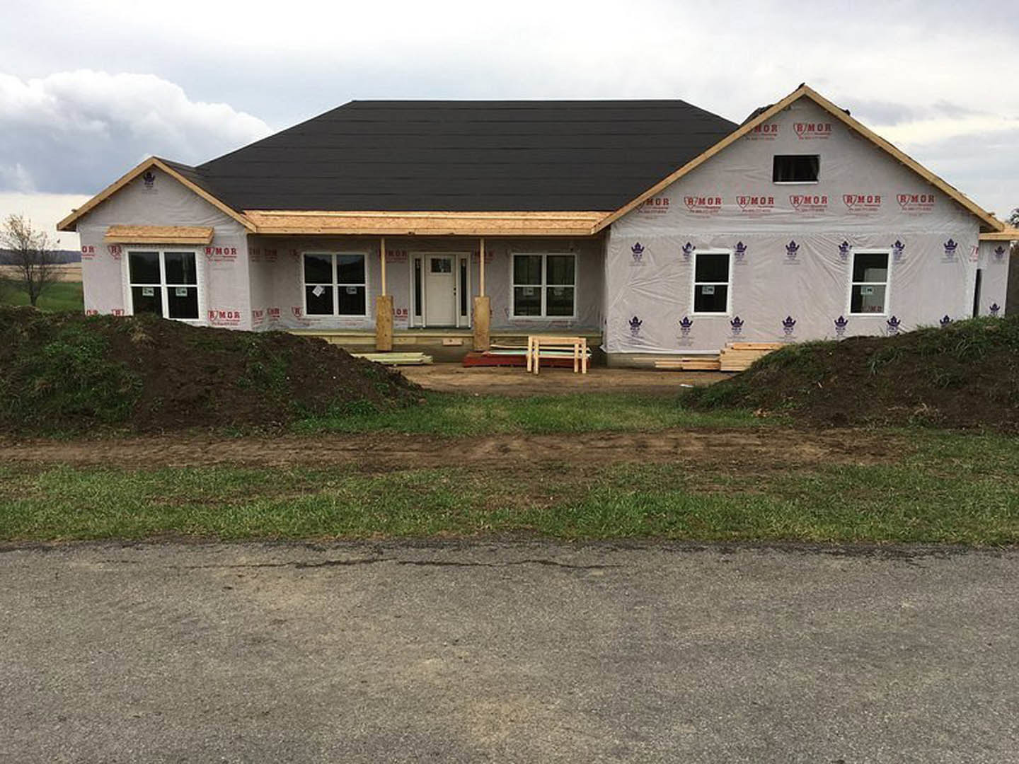 Modern house under construction featuring a black roof, white door with window, white-framed windows, black exterior wall, surrounded by dirt and patches of grass under a cloudy