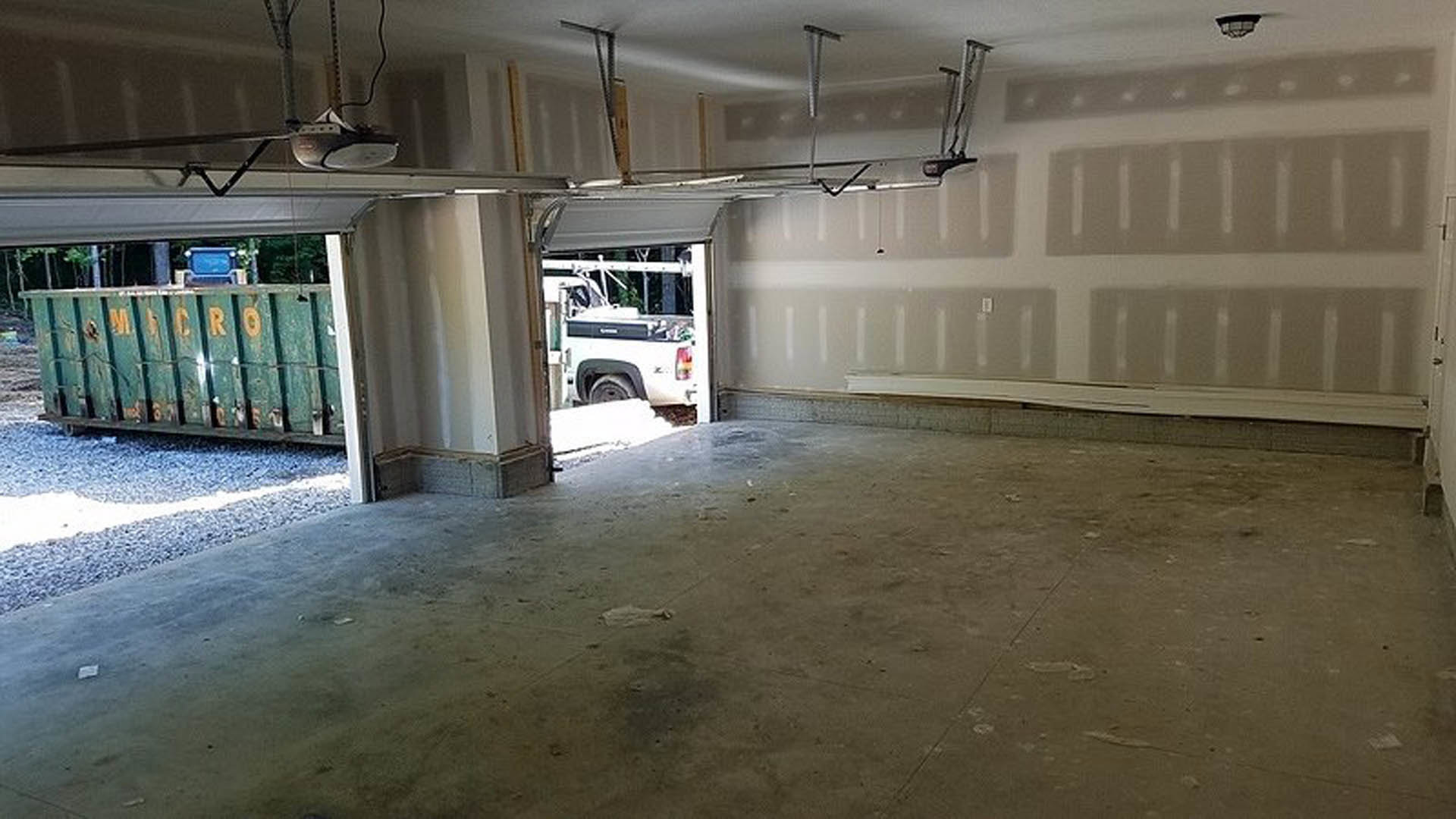 Garage interior with concrete floor, open door, white truck parked in the background, green storage container in foreground, white plaster walls, exposed ceiling beams