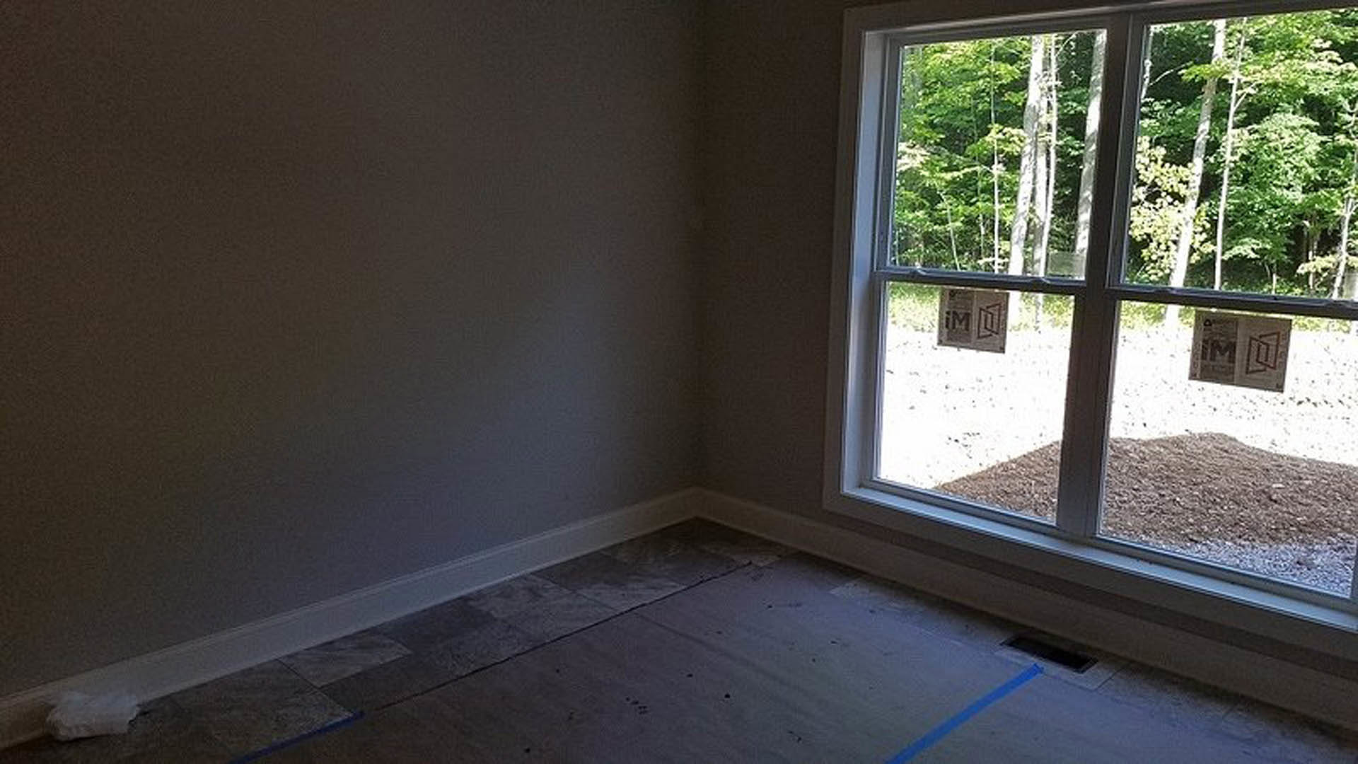 Sunlit room featuring a large window, smooth wood flooring, white walls, and views of greenery outside
