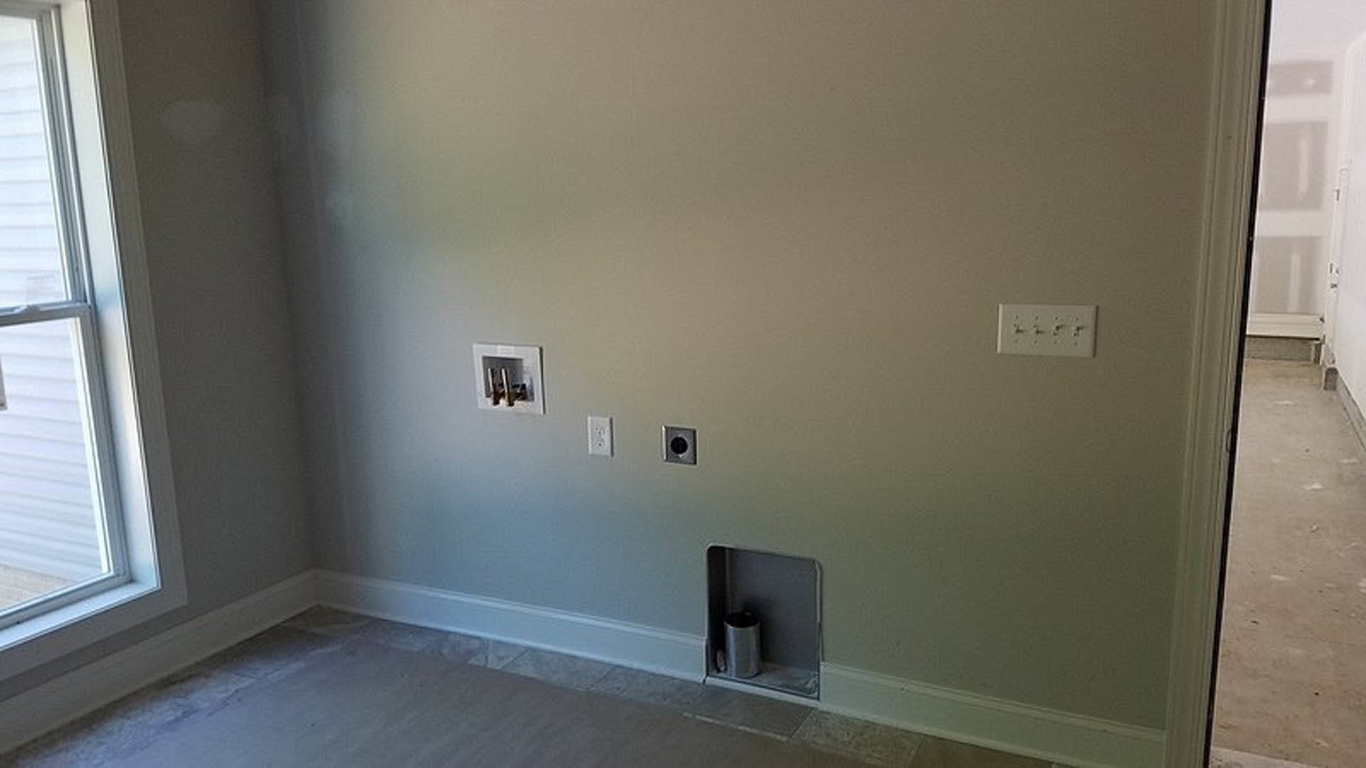 White plaster wall with two electrical outlets, a rectangular hole, and a shelf holding a grey cylindrical object; black circle on square metal plate and scattered black spots