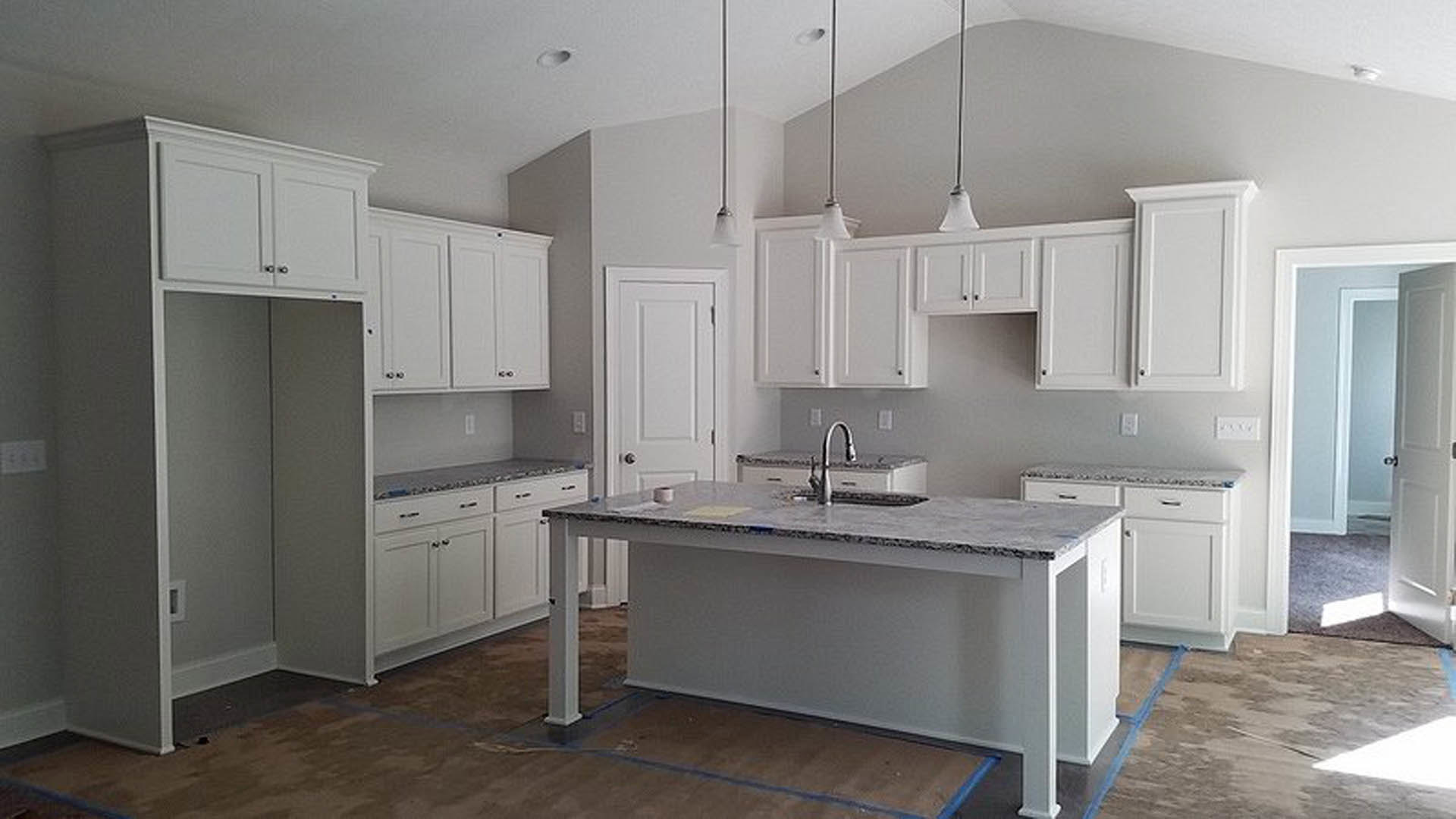 Kitchen with white shaker cabinets, marble countertops, stainless steel sink, blue painter’s tape on counter, light wood flooring, and a white rectangular appliance with black