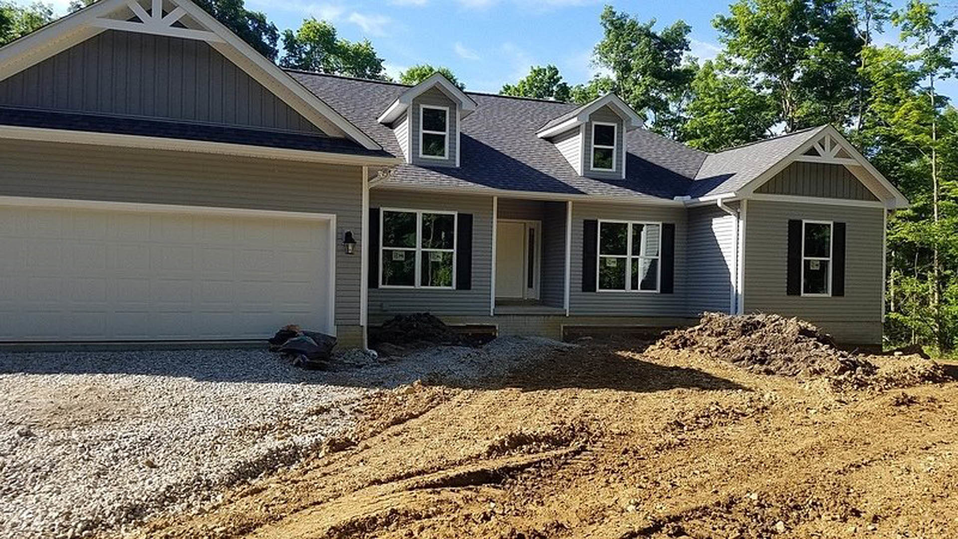 Two-story home with white siding, gray roof, white-framed windows, and a white garage door; concrete driveway leads to the entrance, dirt yard in front, small trees at the edge of