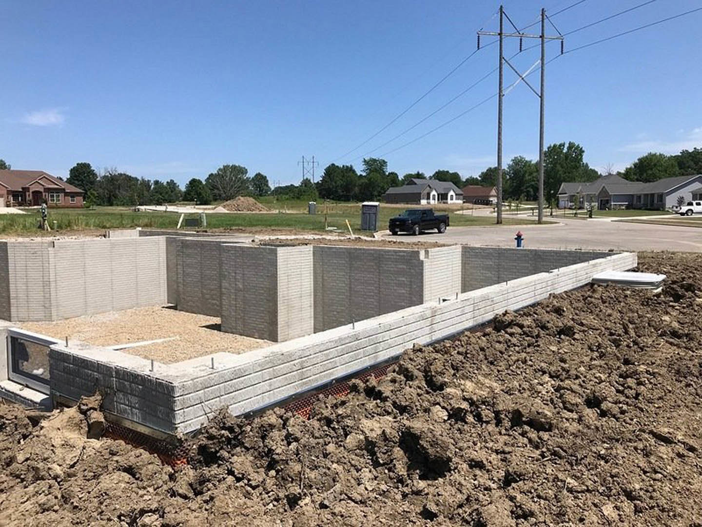 Partially built house with red roof, exposed foundation, dirt construction site, tall power lines, black truck, and blue sky with scattered clouds