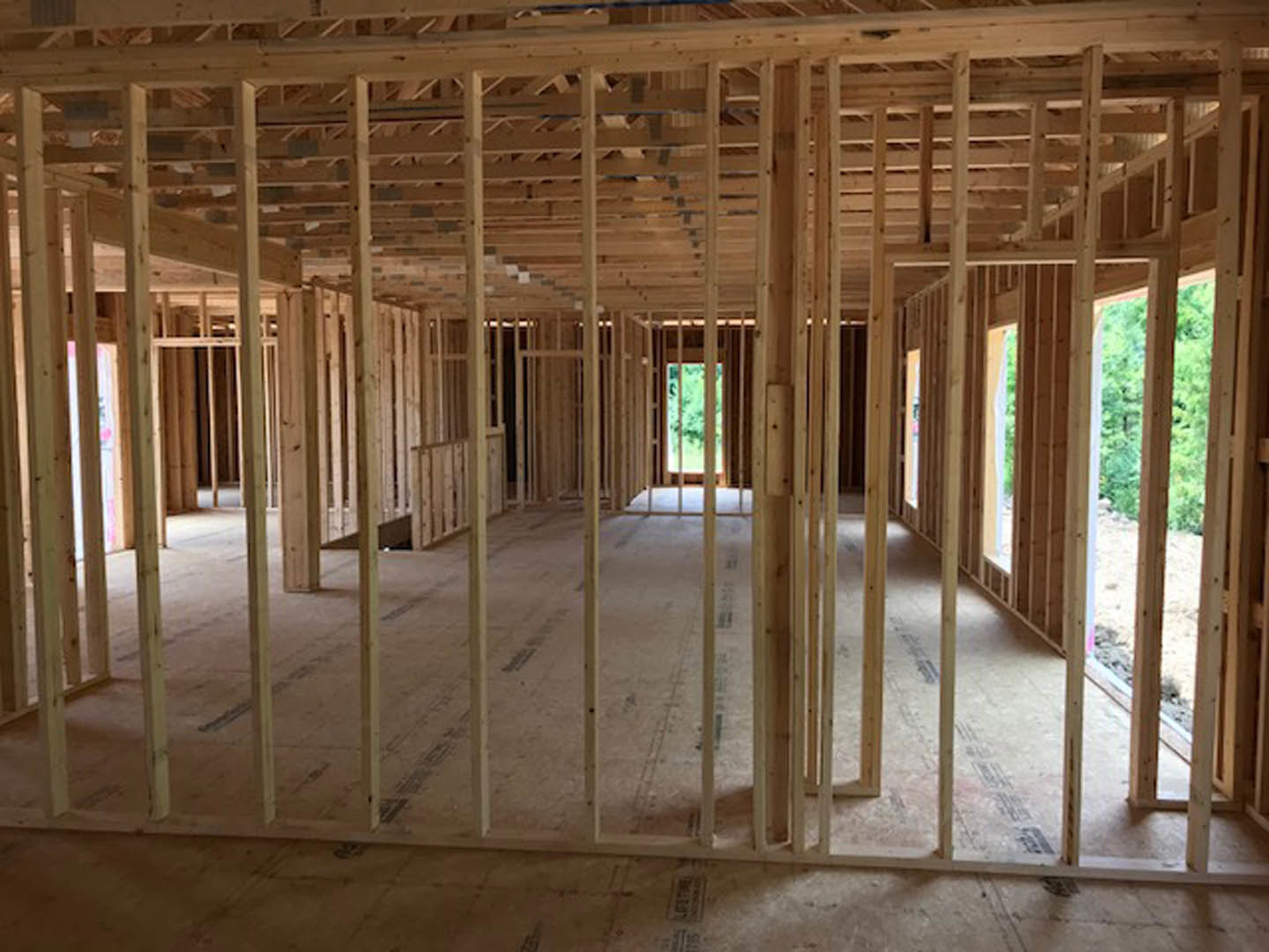 Exposed wood framing and beams inside a partially built house, unfinished floors and open window revealing greenery outside