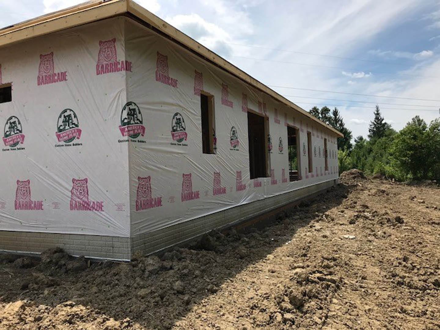 Partially built house with exposed framing, plastic sheeting covering windows, surrounded by dirt field and mature trees under cloudy sky