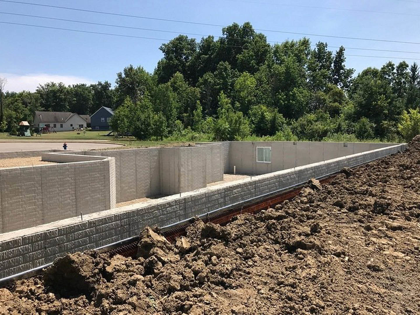 White house under construction with exposed red metal framing, dirt piles in foreground, and surrounding trees with visible power lines.