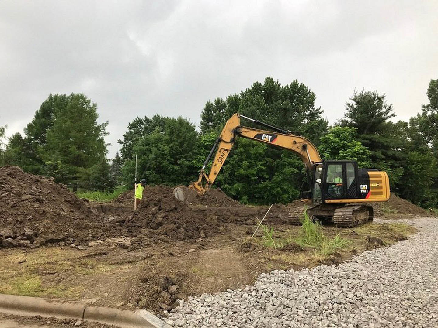Yellow excavator with extended arm digging in dirt beside pile of rocks, surrounded by trees and construction site soil