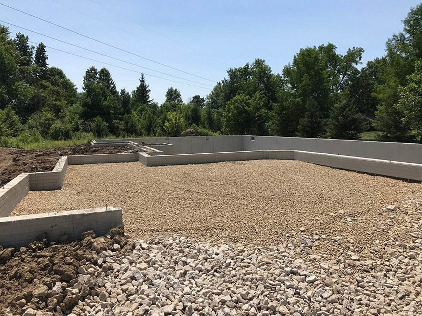 Gravel and rocks scattered across a building site, concrete wall and block in dirt, power lines above green-leaved tree and blue sky
