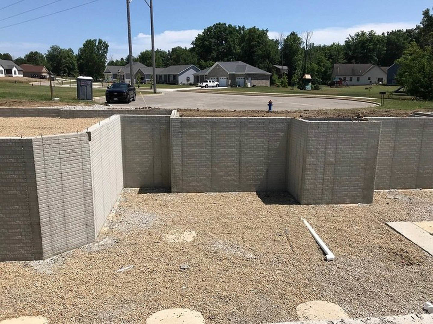 Brick wall with a hole at a residential construction site, dirt ground in foreground, black truck and white car parked near partially built house, black trash can with white lid