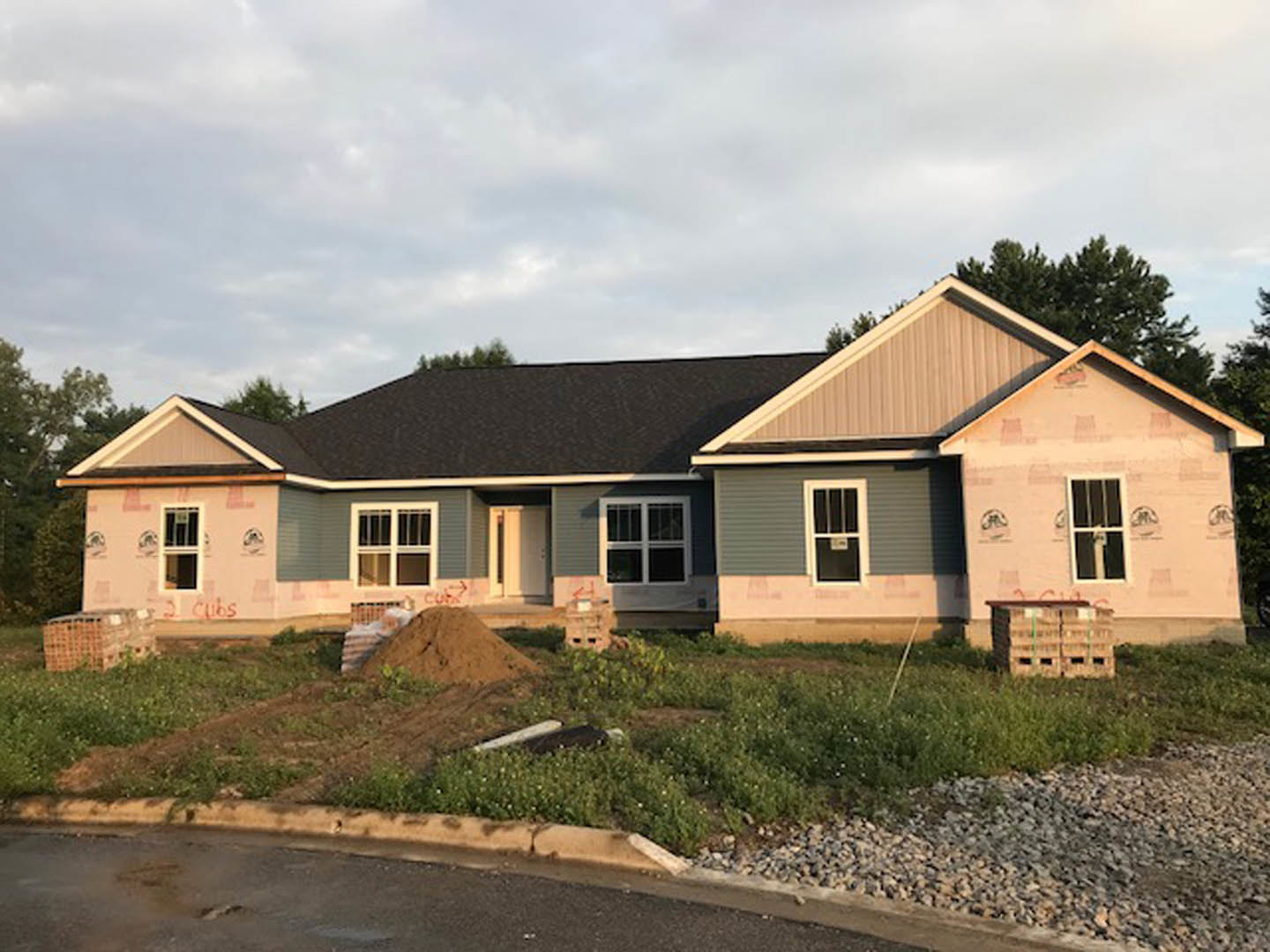 Two-story house under construction with gray roof, white door, white-framed windows, dirt pile in front yard, gravel bed, surrounding trees, and a stack of boxes on a truck nearby