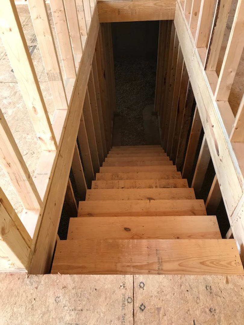 Wooden staircase with matching handrail leading down to a basement, surrounded by neutral walls and flooring, visible wood grain and knots on stair treads.