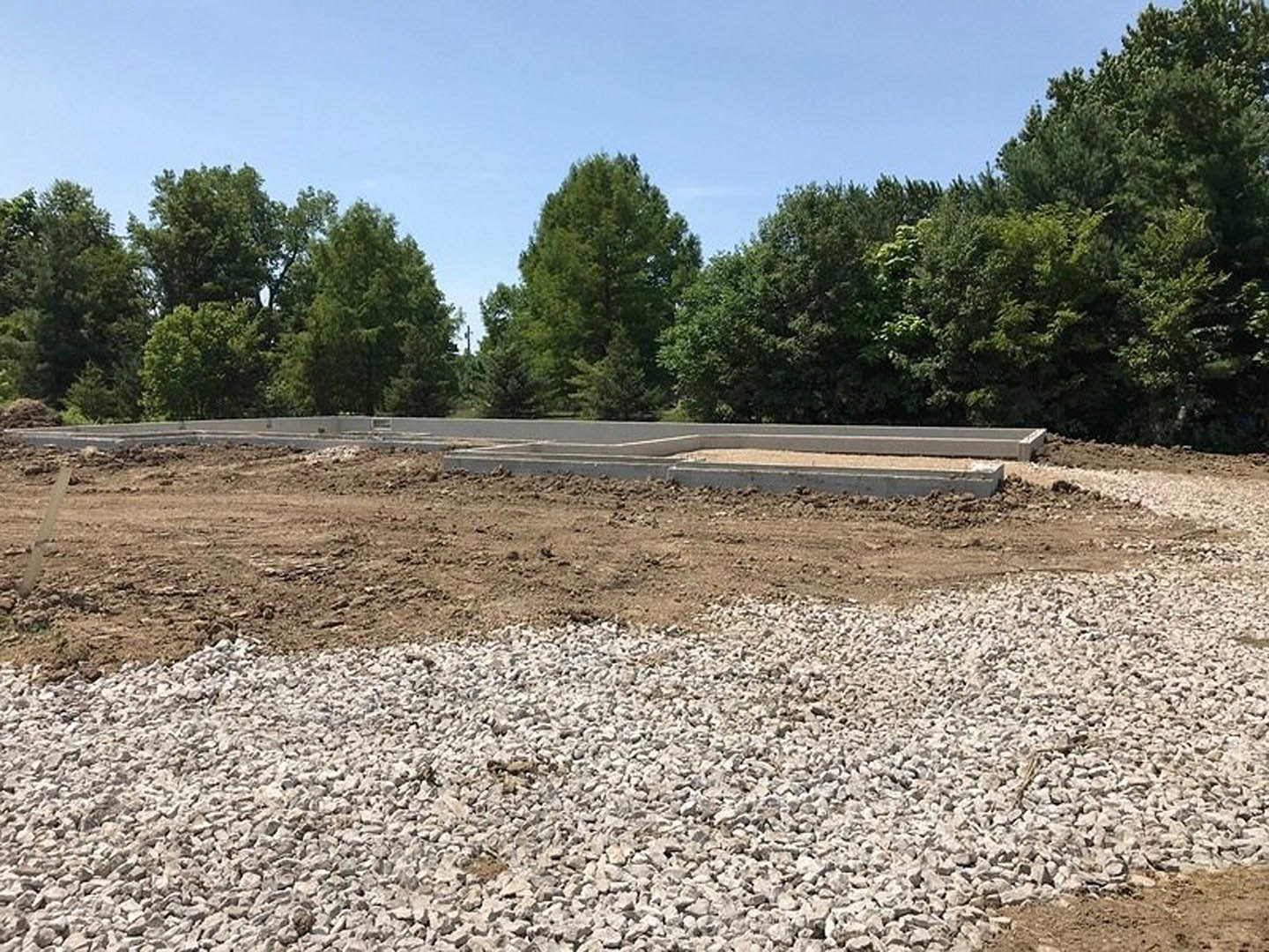 Gravel and dirt landscaping surrounding a modern home, bordered by a concrete curb, with mature trees and blue sky in the background