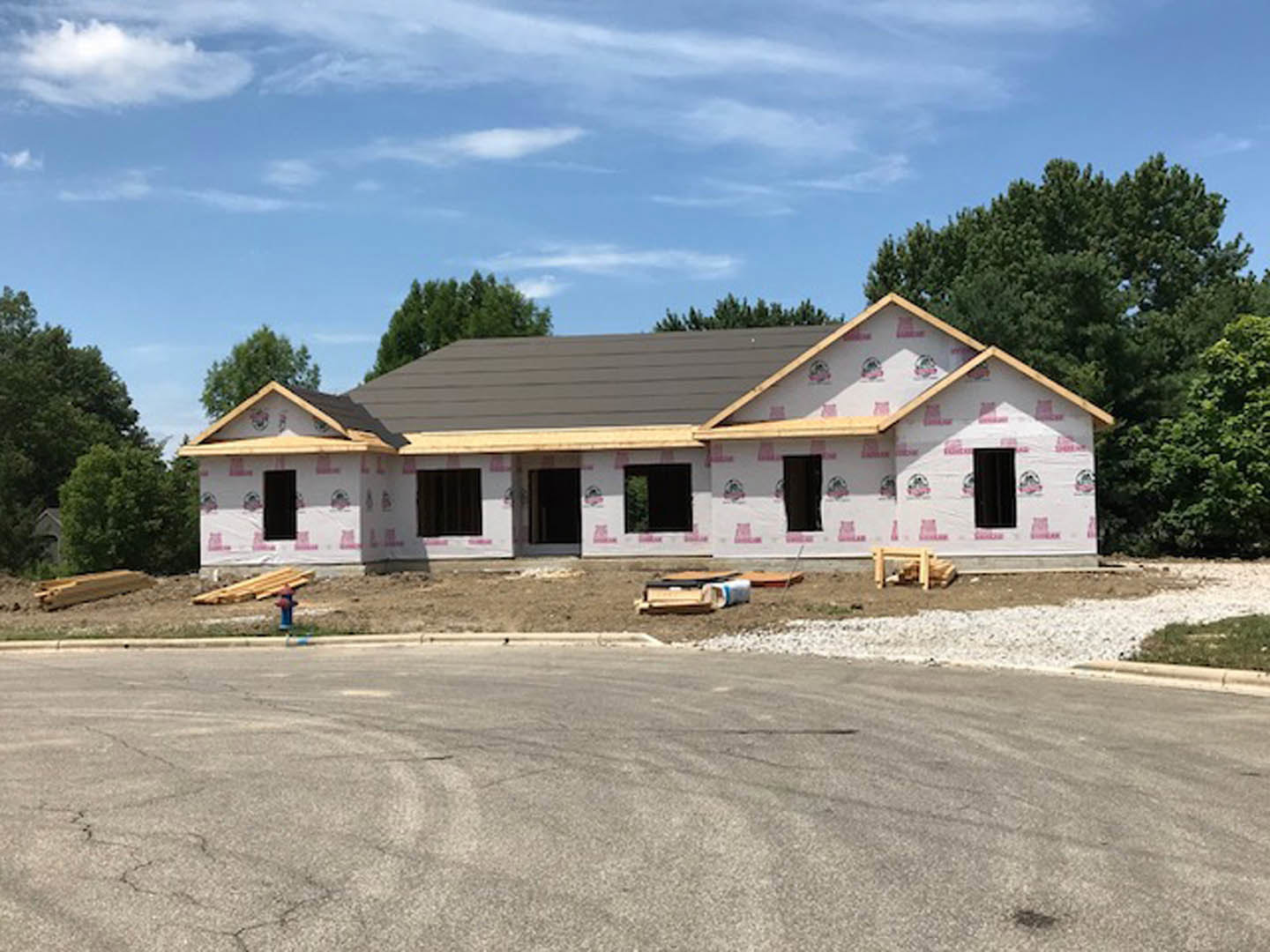 Two-story house under construction with exposed framing, gray roof, and surrounding trees in the background