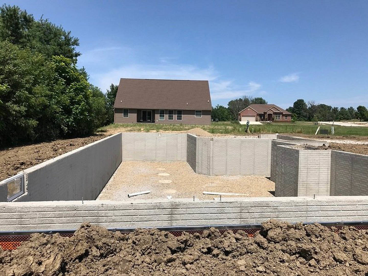 Framed house under construction with brown roof, exposed brick wall, pile of dirt near foundation, mature trees and blue sky in background