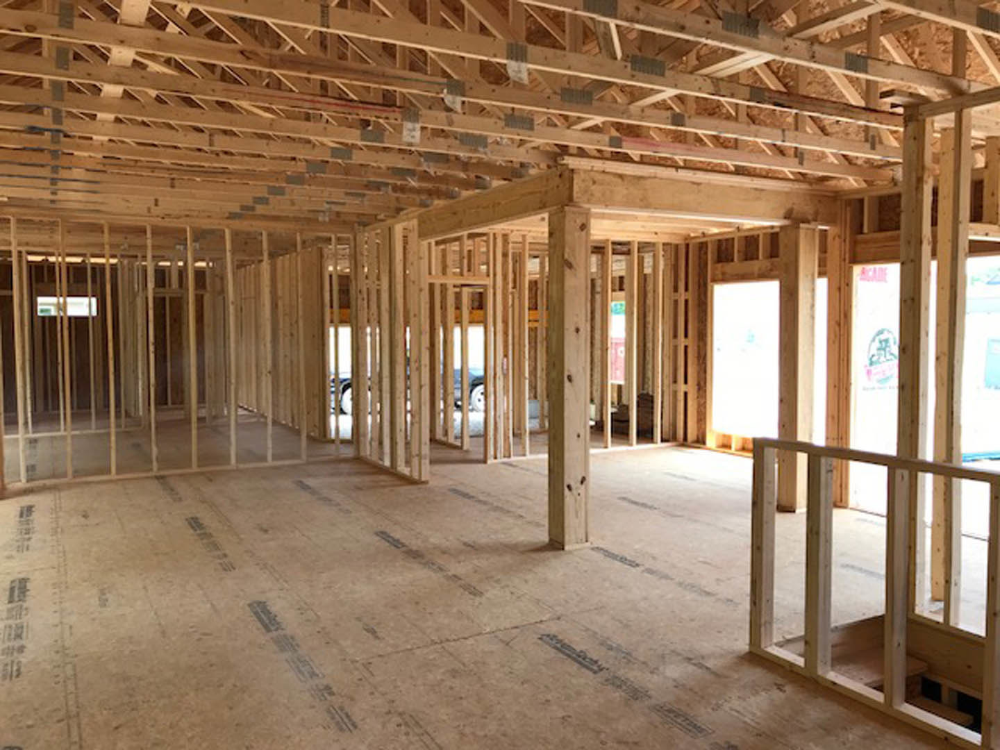 Wood-framed house under construction with exposed beams, unfinished flooring marked with black tape, and a white board leaning against the wall.