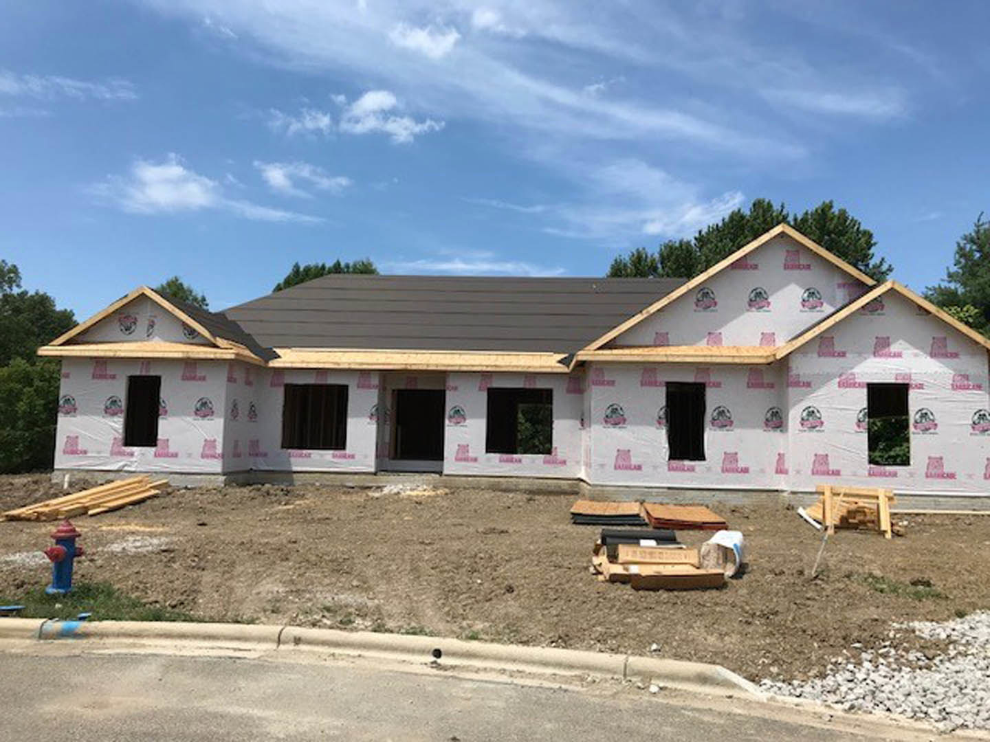 Framed house under construction with exposed wooden beams, stacked wood pallets in foreground, partially finished roof, cloudy sky overhead