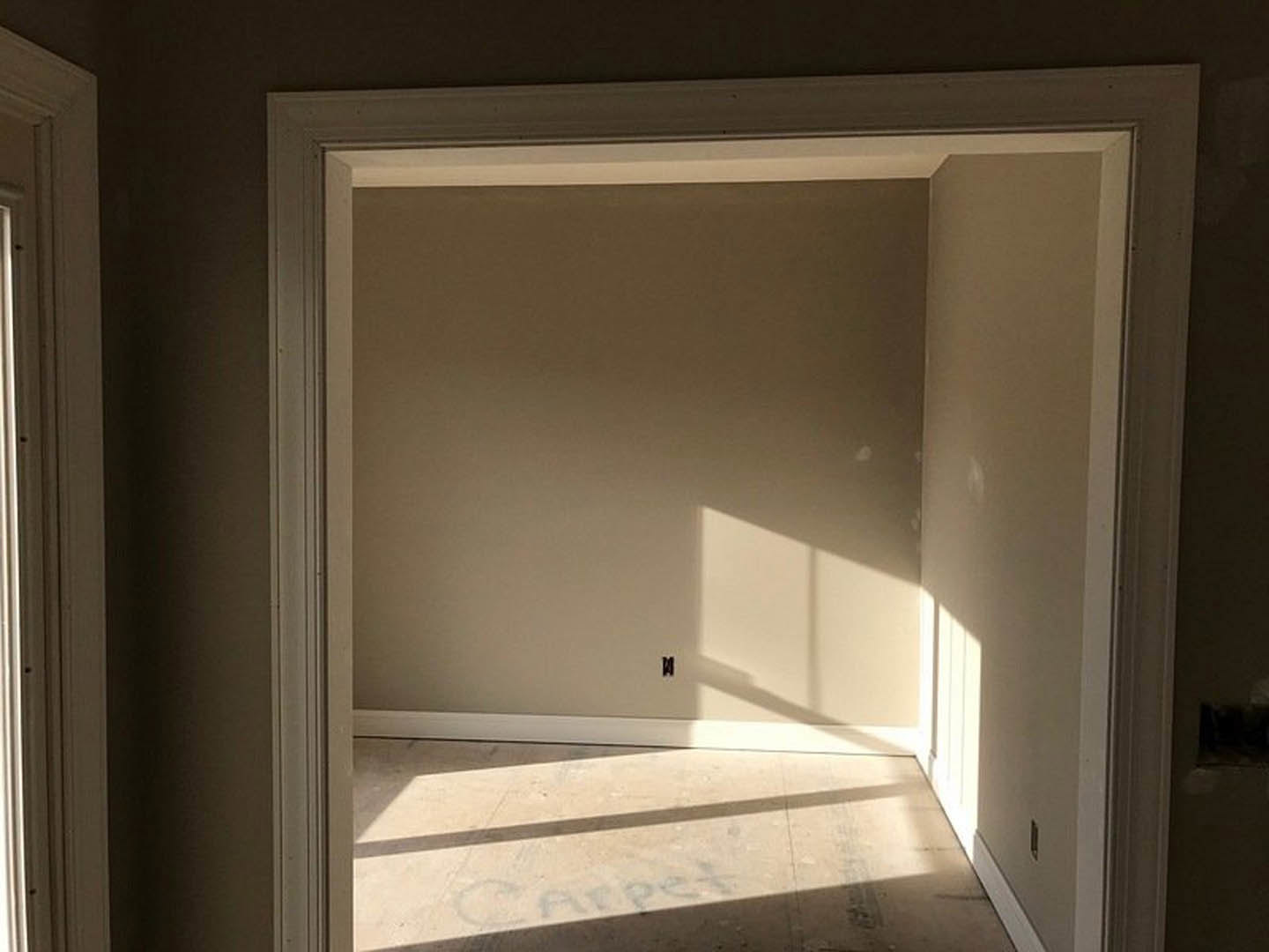 White plaster walls and door frame in a sunlit room, with shadows cast on the floor and wall, and a window visible in the background.