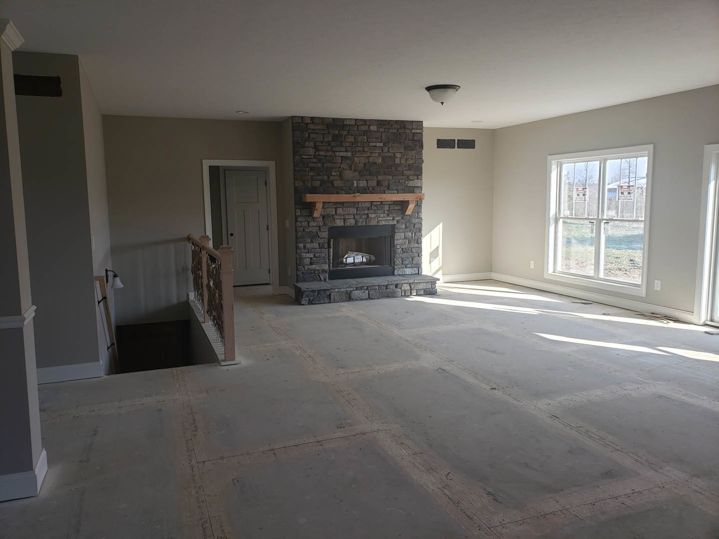 Living room with stone fireplace, wood mantel shelf, light fixture on wall, large window with signs, white door with child, black storage box with bag, plaster walls, hardwood