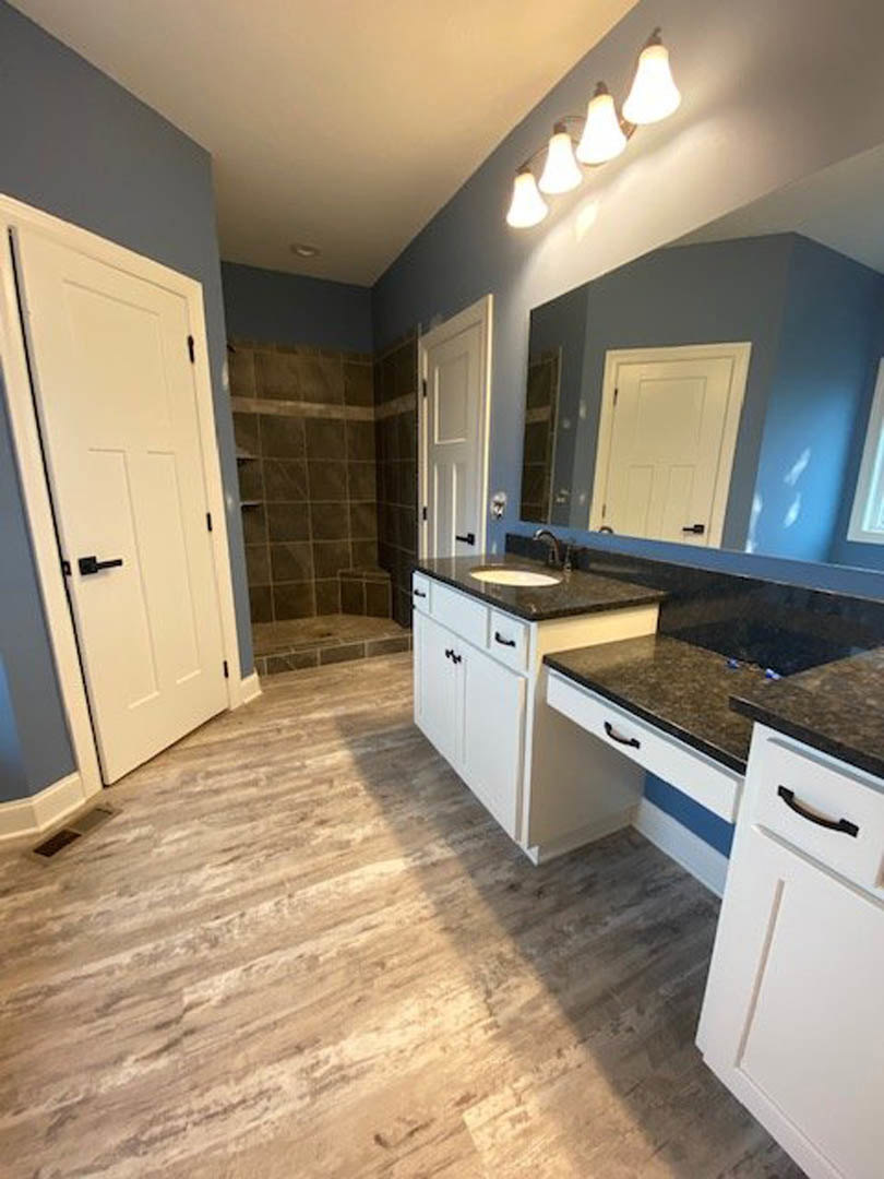 Bathroom featuring wood flooring, white countertop with integrated sink, row of modern light fixtures above mirror, glass-enclosed shower, white cabinetry, blue accent wall, and
