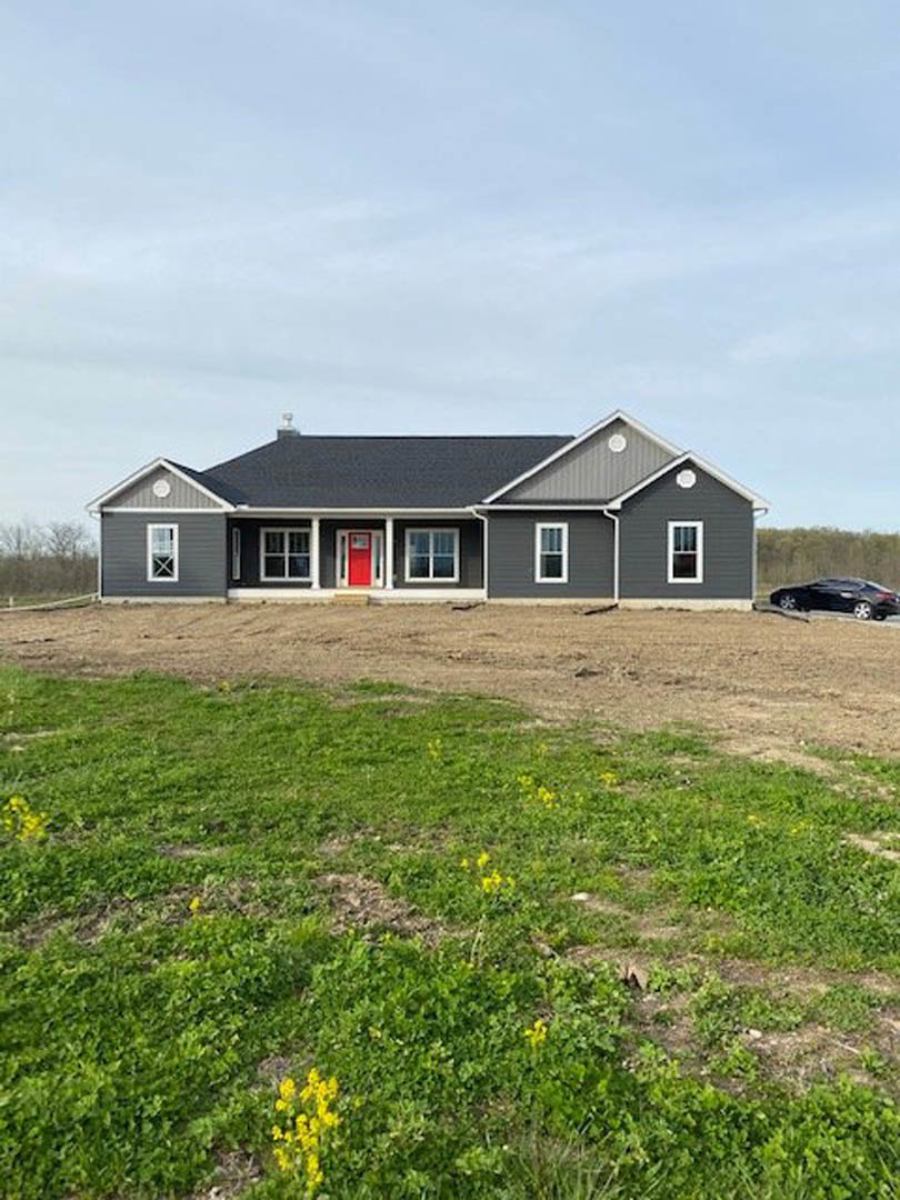 White farmhouse with gray roof, red front door, and large windows, surrounded by grassy field with patches of dirt; yellow flowers in foreground, parked car partially visible in