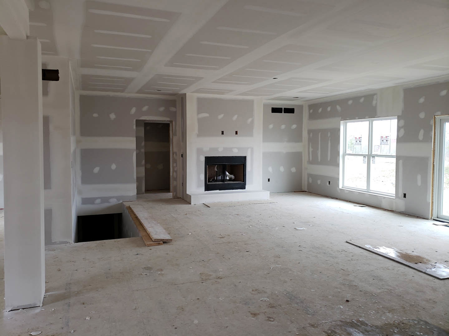 Living room with black fireplace, wood flooring, plaster walls, large window displaying signs, and white decorative object inside fireplace