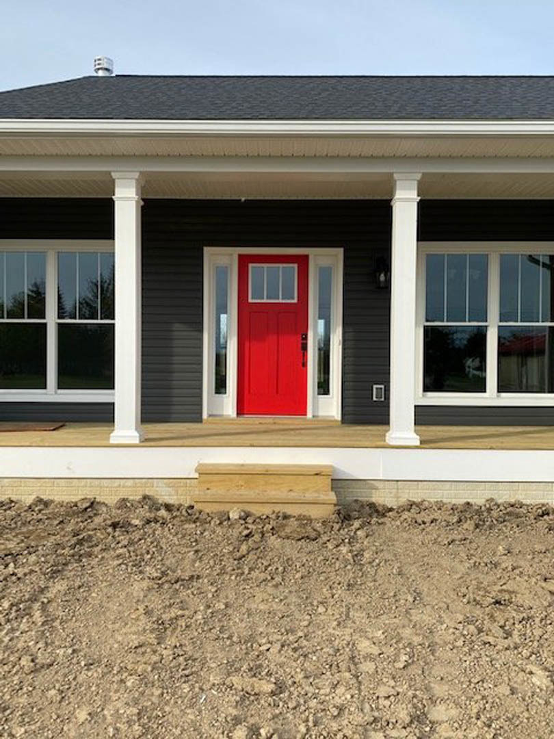 Red front door with glass window set in wood-paneled exterior, white trim, and adjacent window; dirt ground and roof visible.