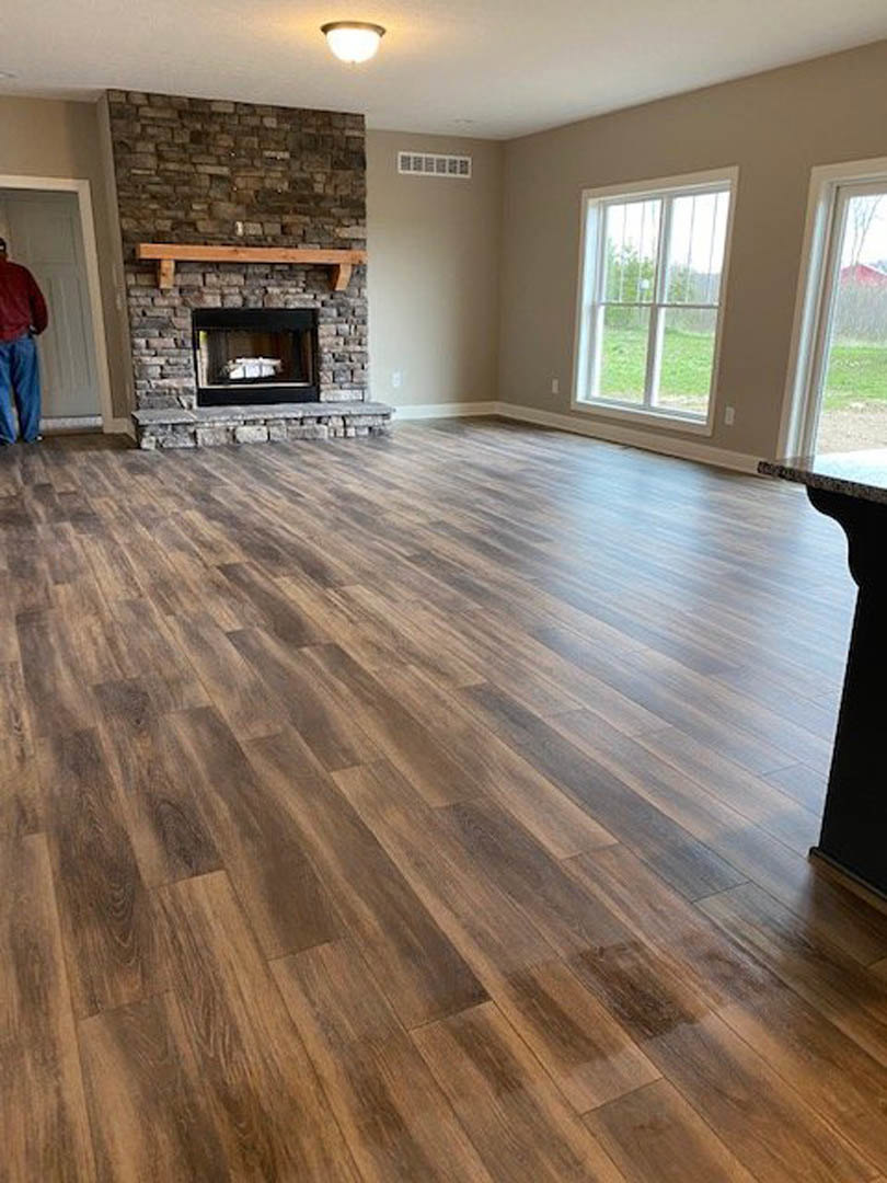 Living room with hardwood floors, stone fireplace, wall-mounted light fixture, large window overlooking green lawn, man in red shirt standing by window
