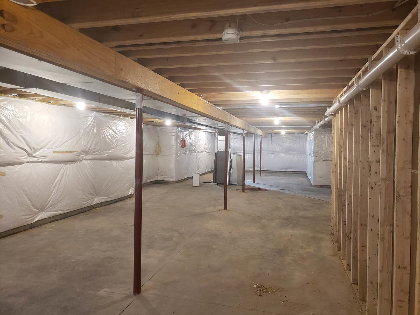 White plaster wall and exposed wooden ceiling beams, concrete floor with metal support pole, white smoke detector mounted on ceiling, plastic coverings draped along walls.