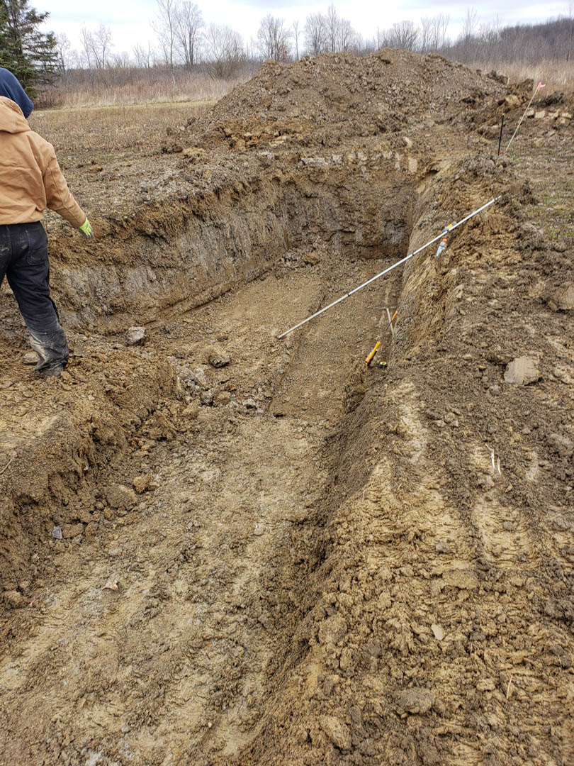 Man in brown jacket and gloves standing in a dirt pit with exposed soil and rocks, pipe visible in hole, outdoor setting with trees and sky in background