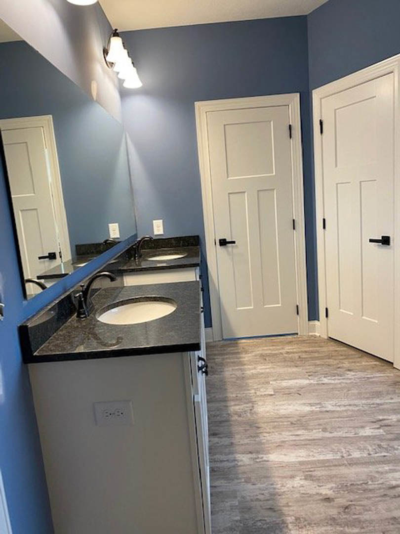 Bathroom with double sinks set in a stone countertop, white cabinetry, wood flooring, open white door with black handle, wall-mounted light fixture, and visible wall outlet.