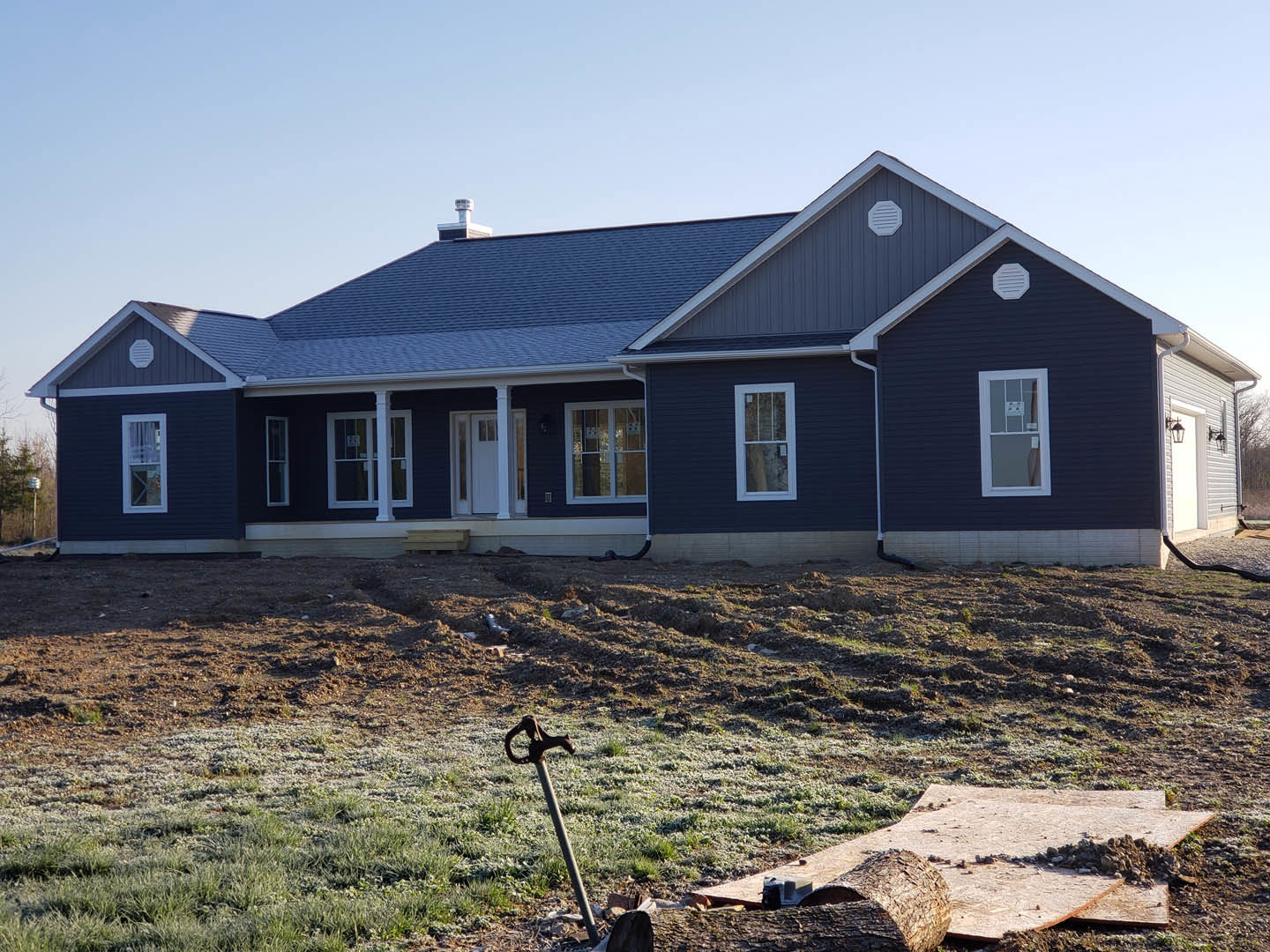 Partially built house with blue roof, white framed windows, exposed siding, surrounded by dirt field and scattered construction materials