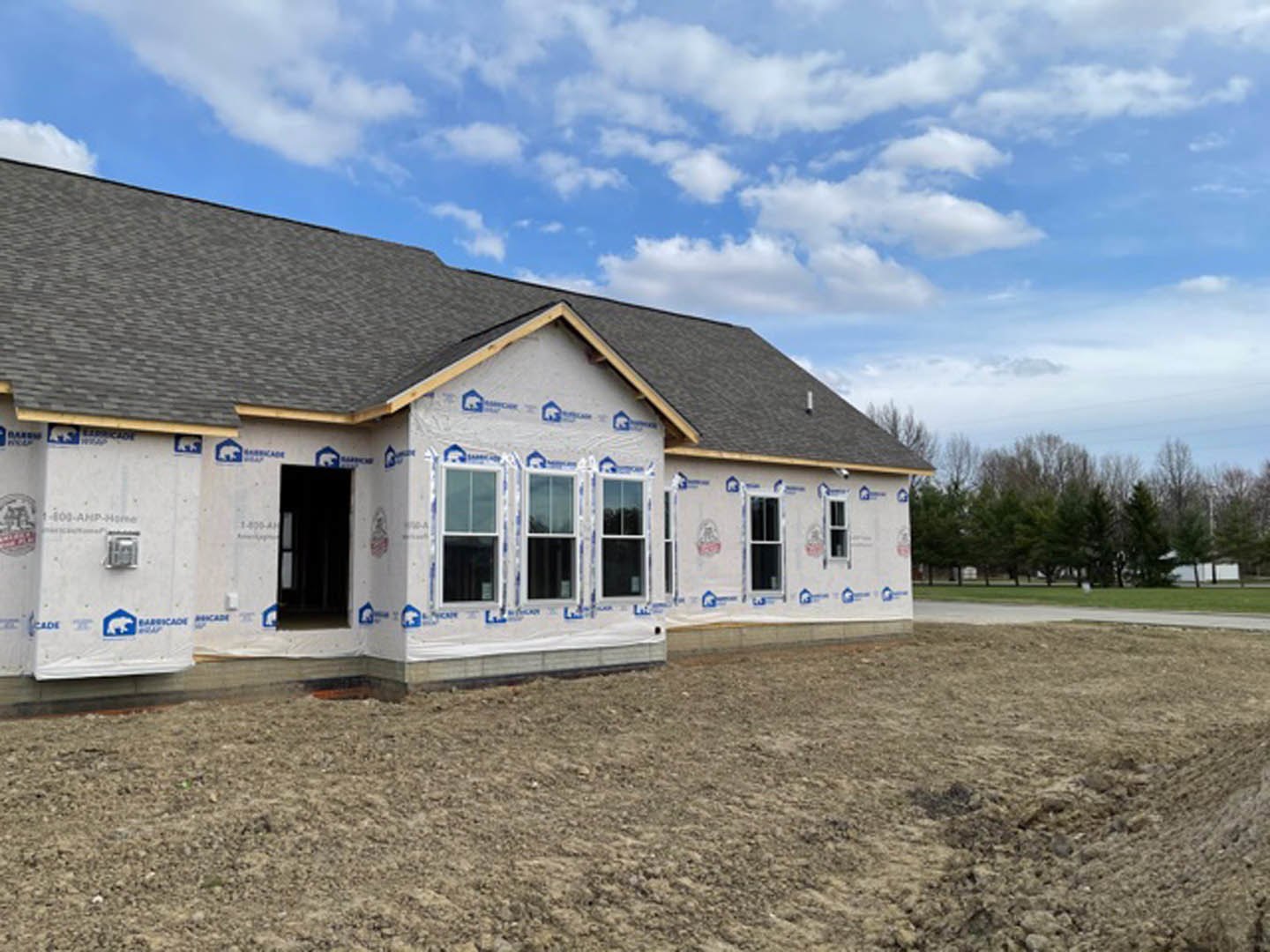 Two-story house under construction with white-framed windows, black and white doors, exposed dirt yard, scattered white construction materials, surrounded by trees under blue sky