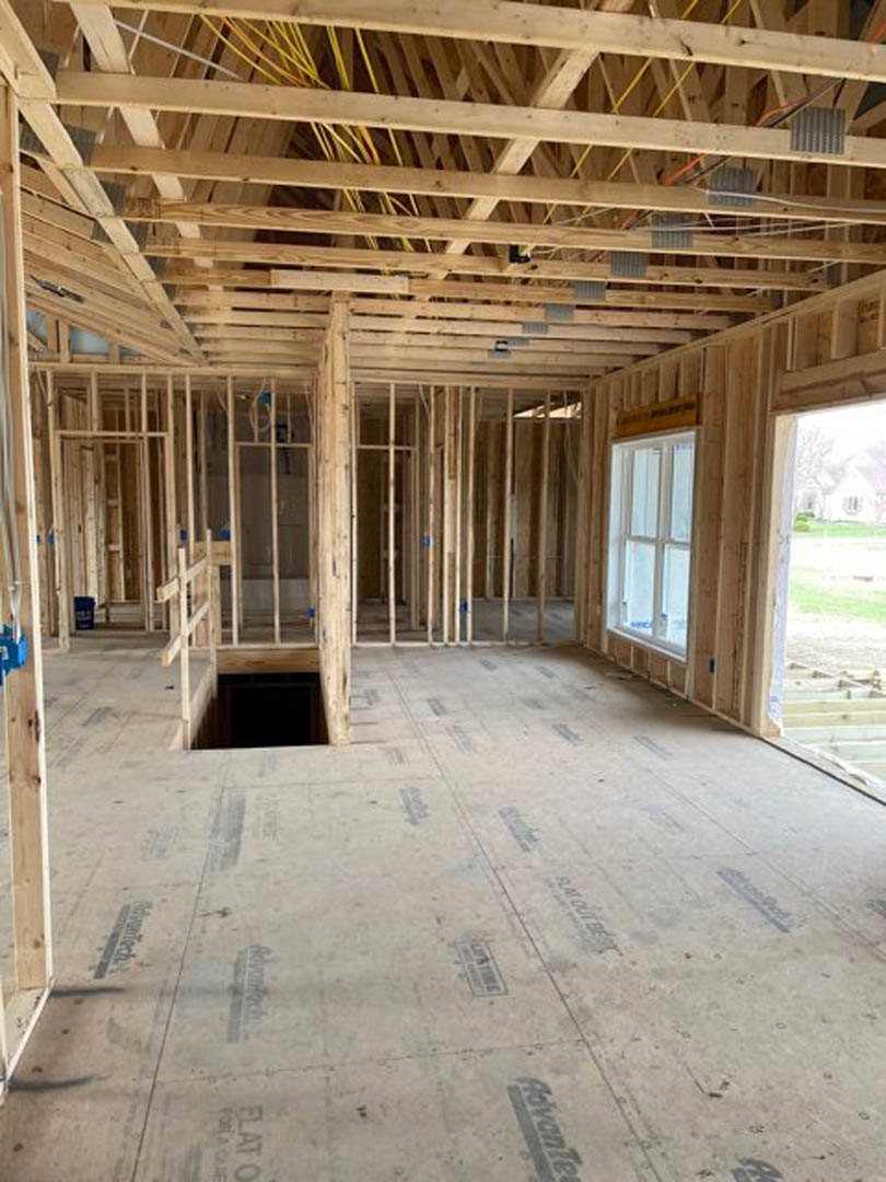 Unfinished room featuring exposed wooden ceiling beams, visible wiring, construction flooring, and a window overlooking grass and an adjacent building