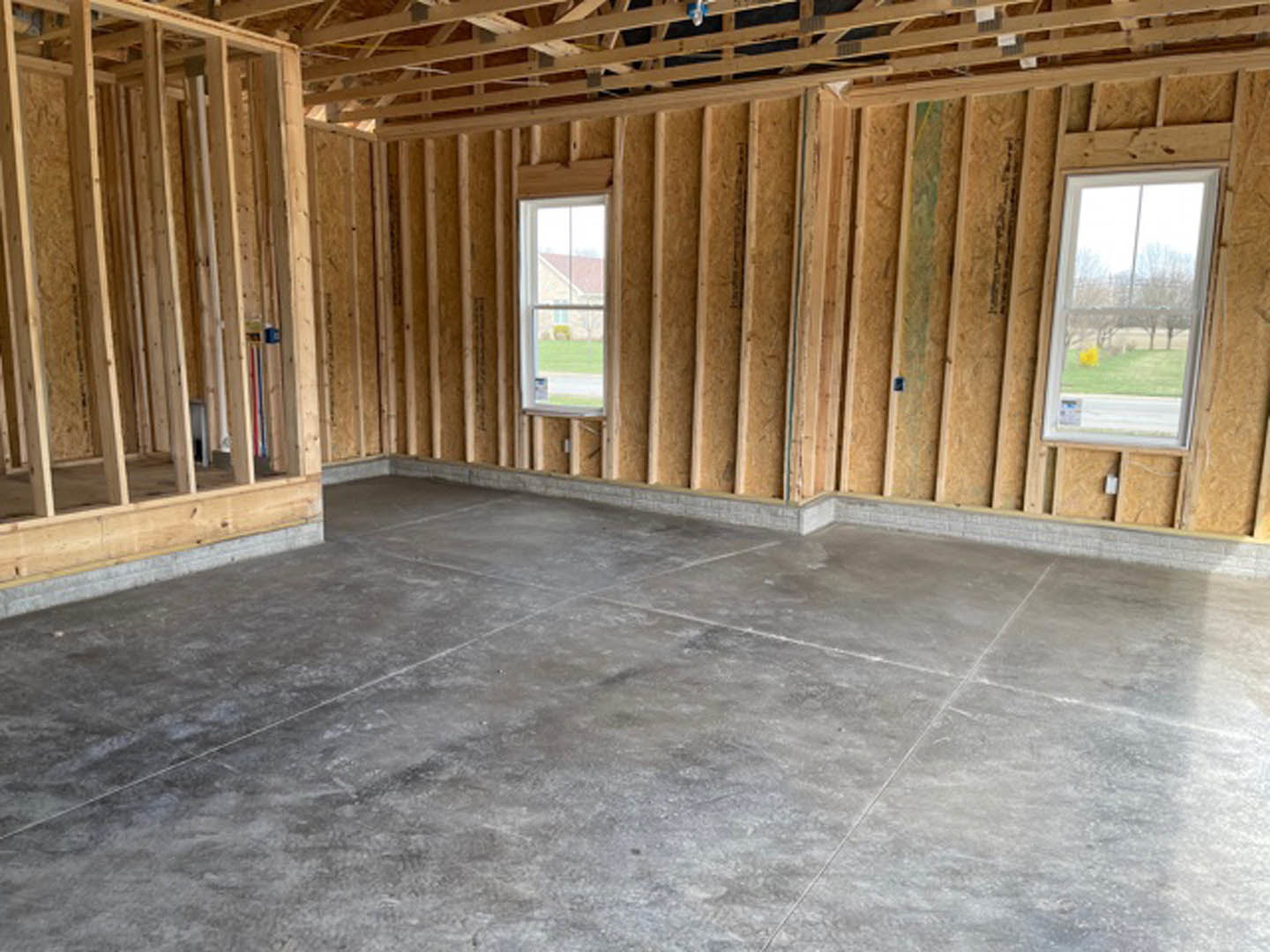 Open living space with exposed wood ceiling beams, large windows overlooking a green lawn, grey concrete flooring, and visible wooden framing around windows and ceiling