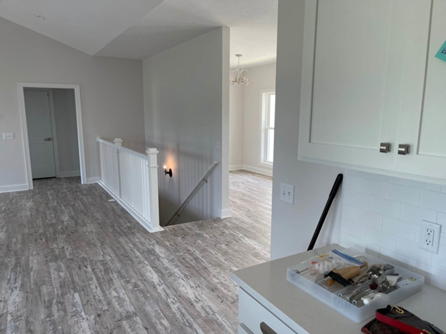 White kitchen with wood laminate flooring, white cabinetry with silver handles, white countertop, and a white door with a silver handle; plastic container with tools and a bag