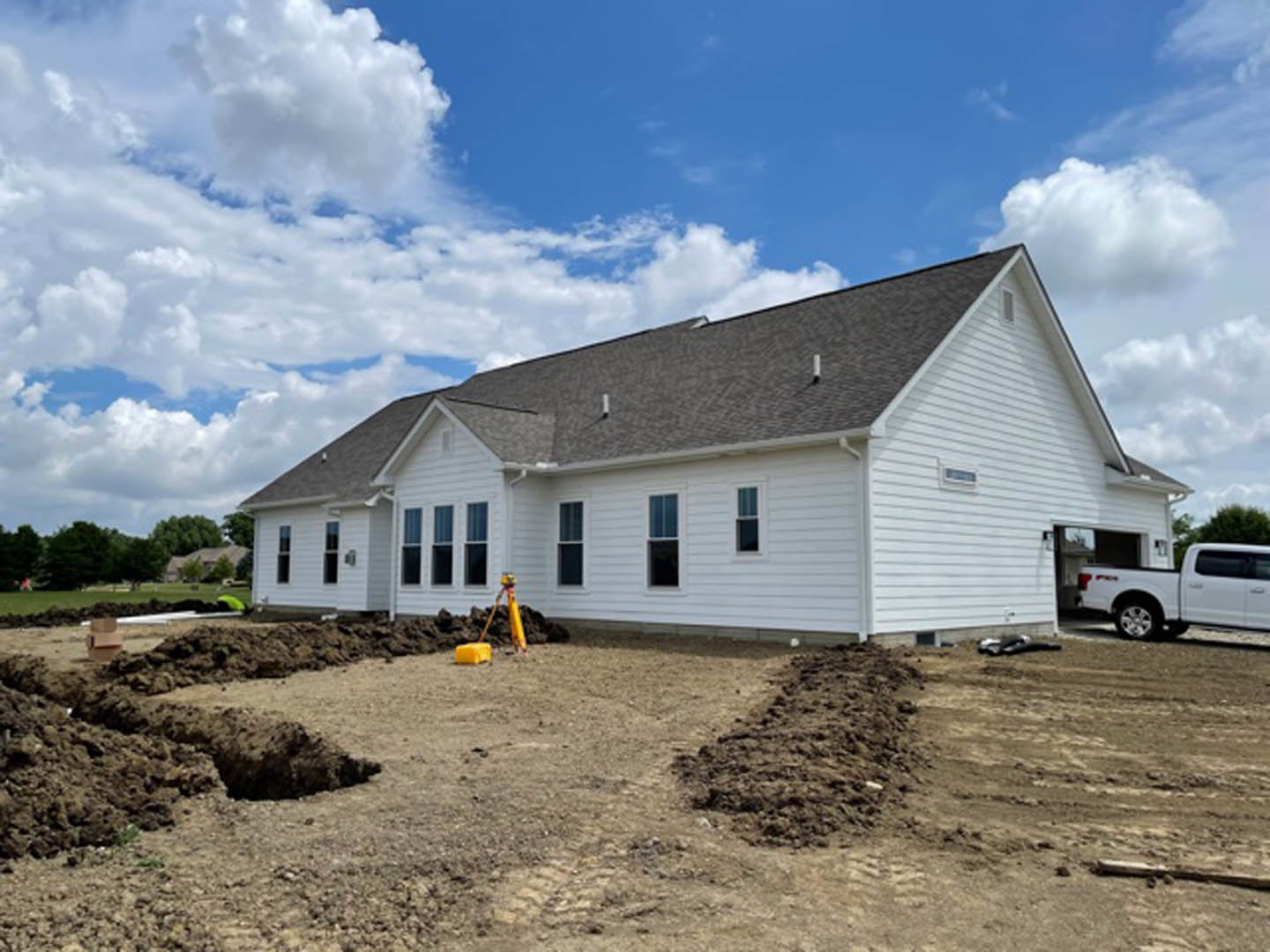 Framed house under construction with exposed wood, dirt piles in foreground, white truck parked near foundation, yellow construction equipment, blue sky with scattered clouds