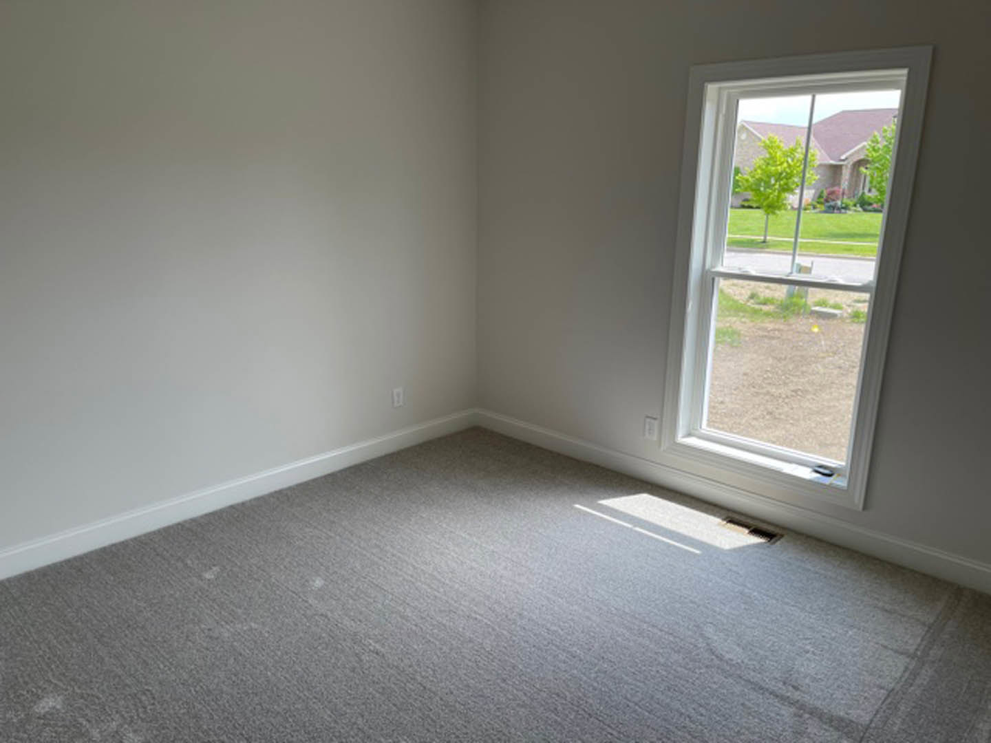 Carpeted room with white plaster walls, large window overlooking a house and grassy yard, natural daylight streaming in