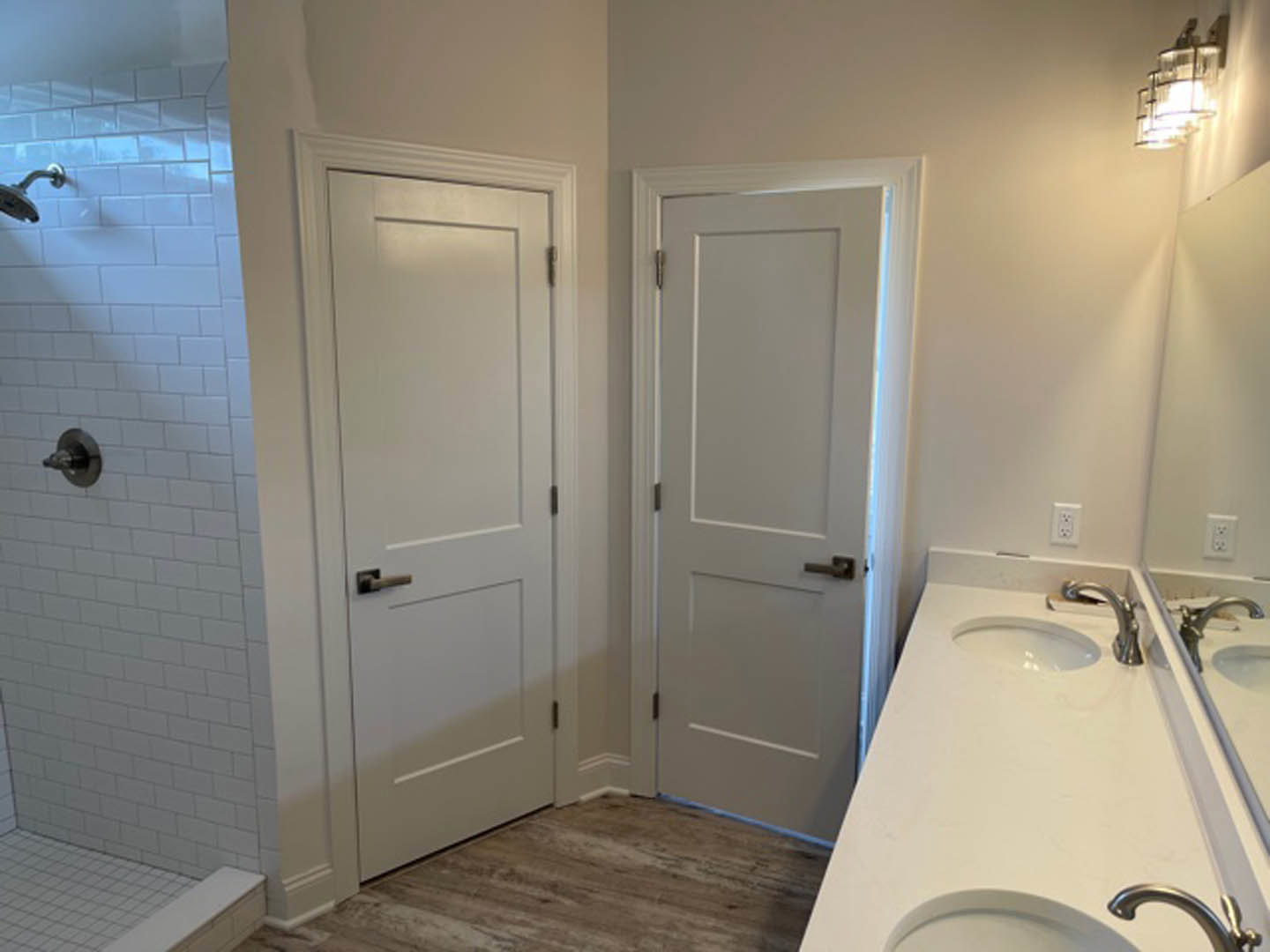 Bathroom with dual white doors, white countertop featuring two sinks and silver faucets, wall-mounted light fixture above mirrors, tiled floor and walls